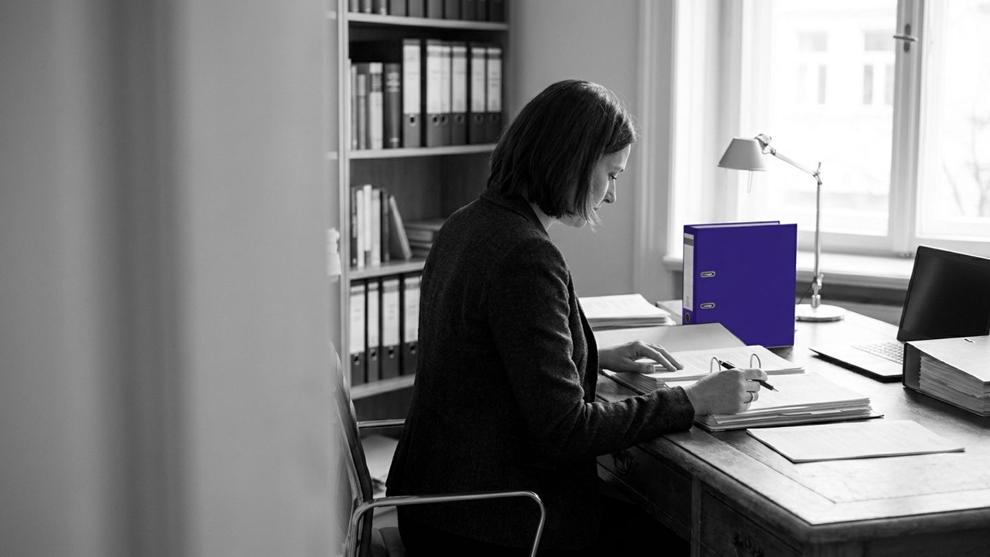 Compliance lead reviewing technical documents and binders at a desk, with a single indigo file spine