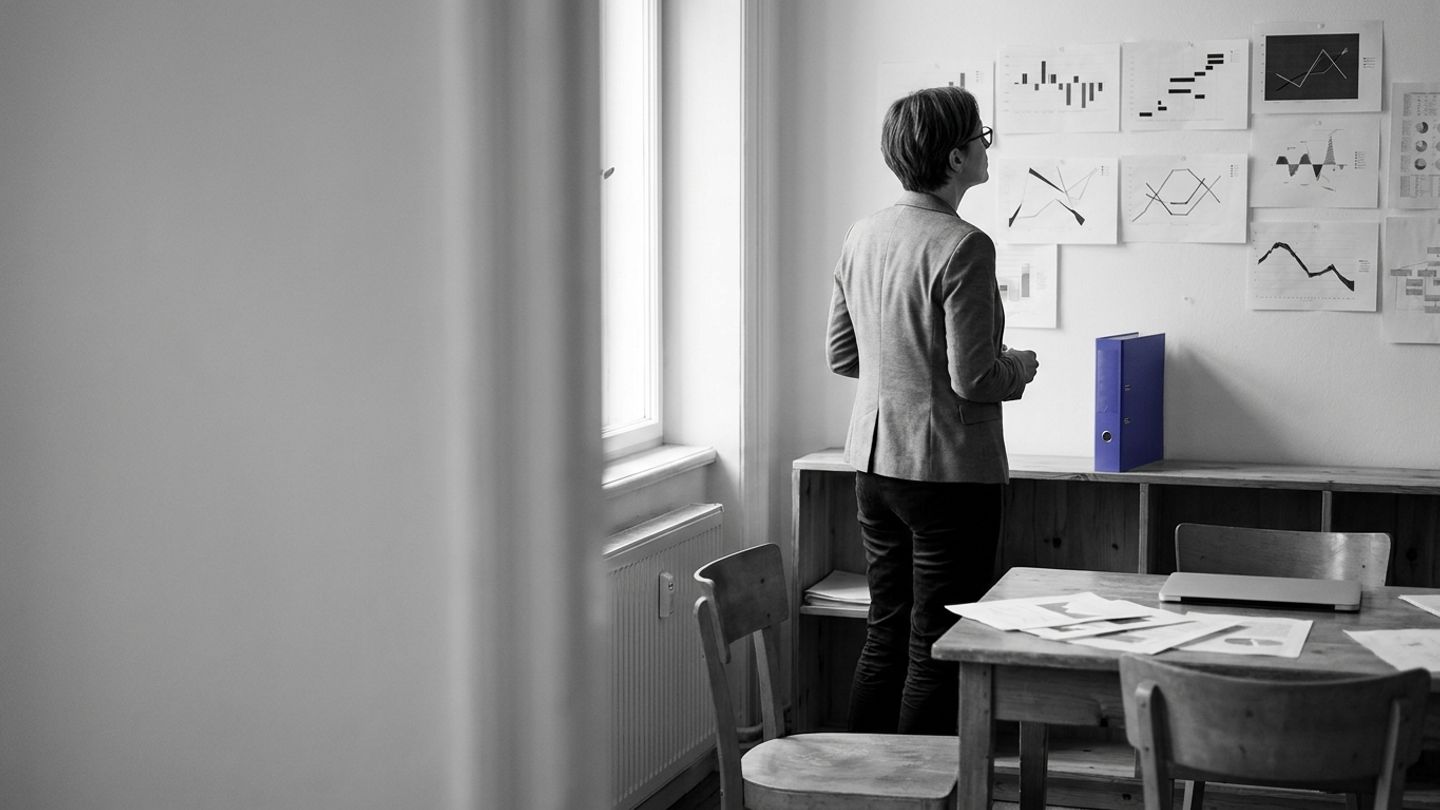 Policy lead studying a wall of pinned documents, with an indigo folder spine on a shelf