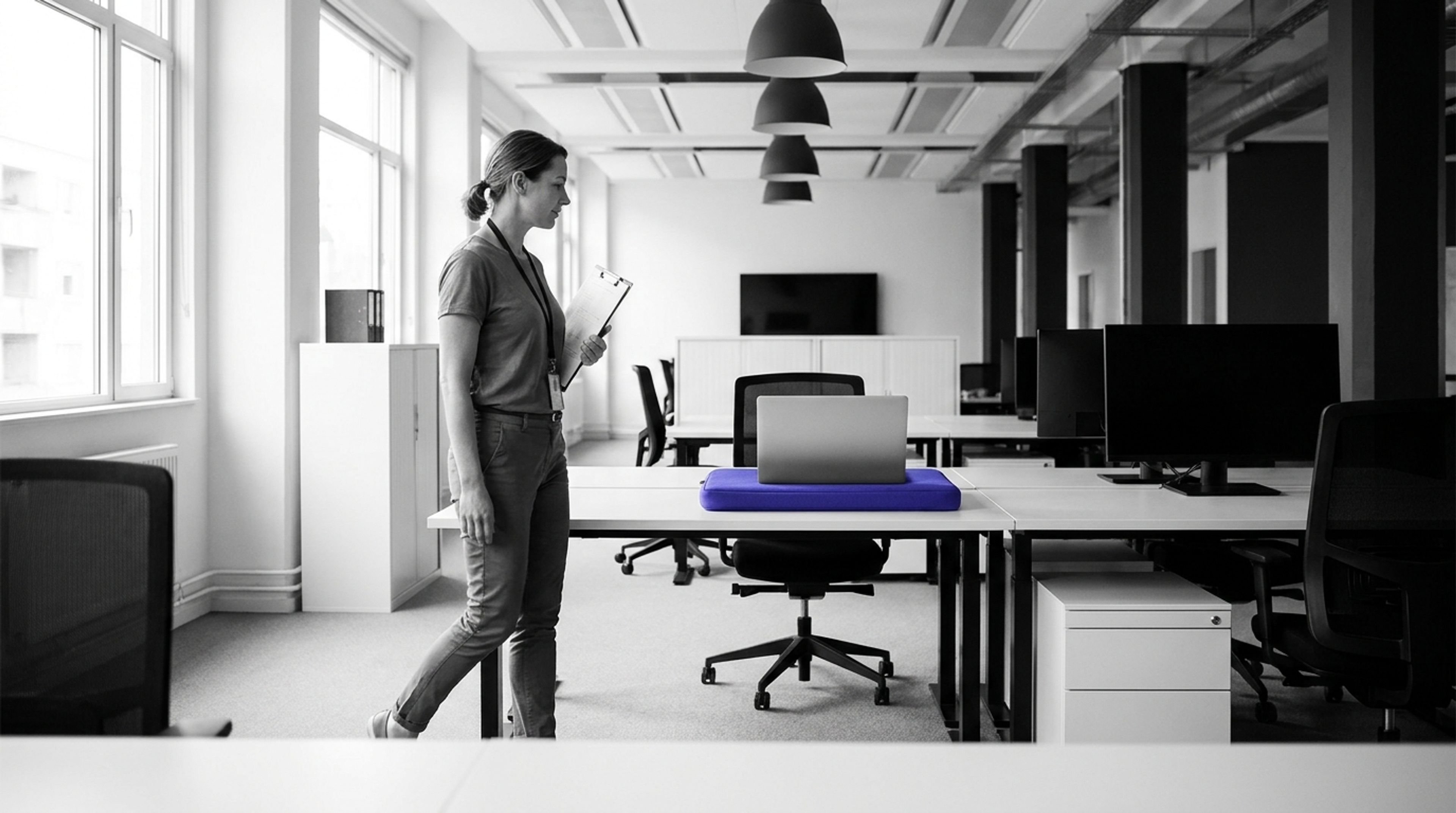 An IT asset manager with a clipboard passes a desk with an indigo laptop sleeve.