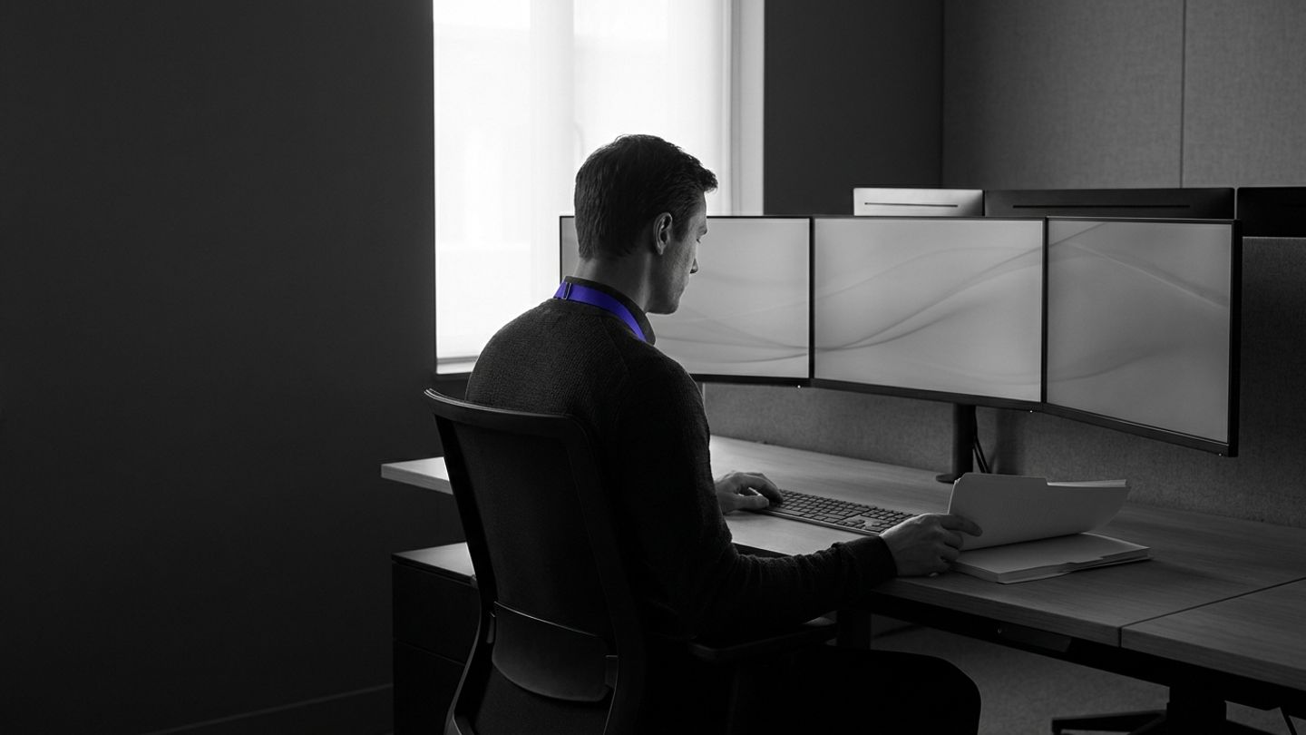 Compliance officer at a desk of monitors, monochrome with an indigo lanyard