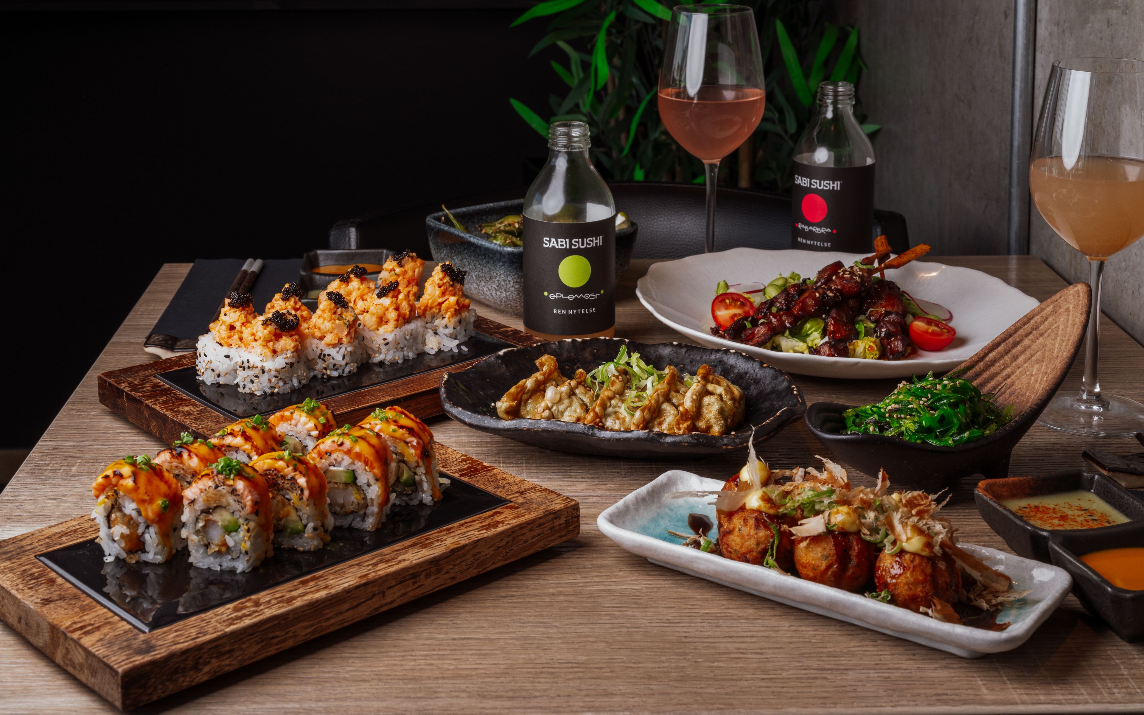 Et bord dekket med japansk mat fra Sabi Sushi, inkludert sushi, gyoza, takoyaki og sjøgressalat, med to glass rosévin.