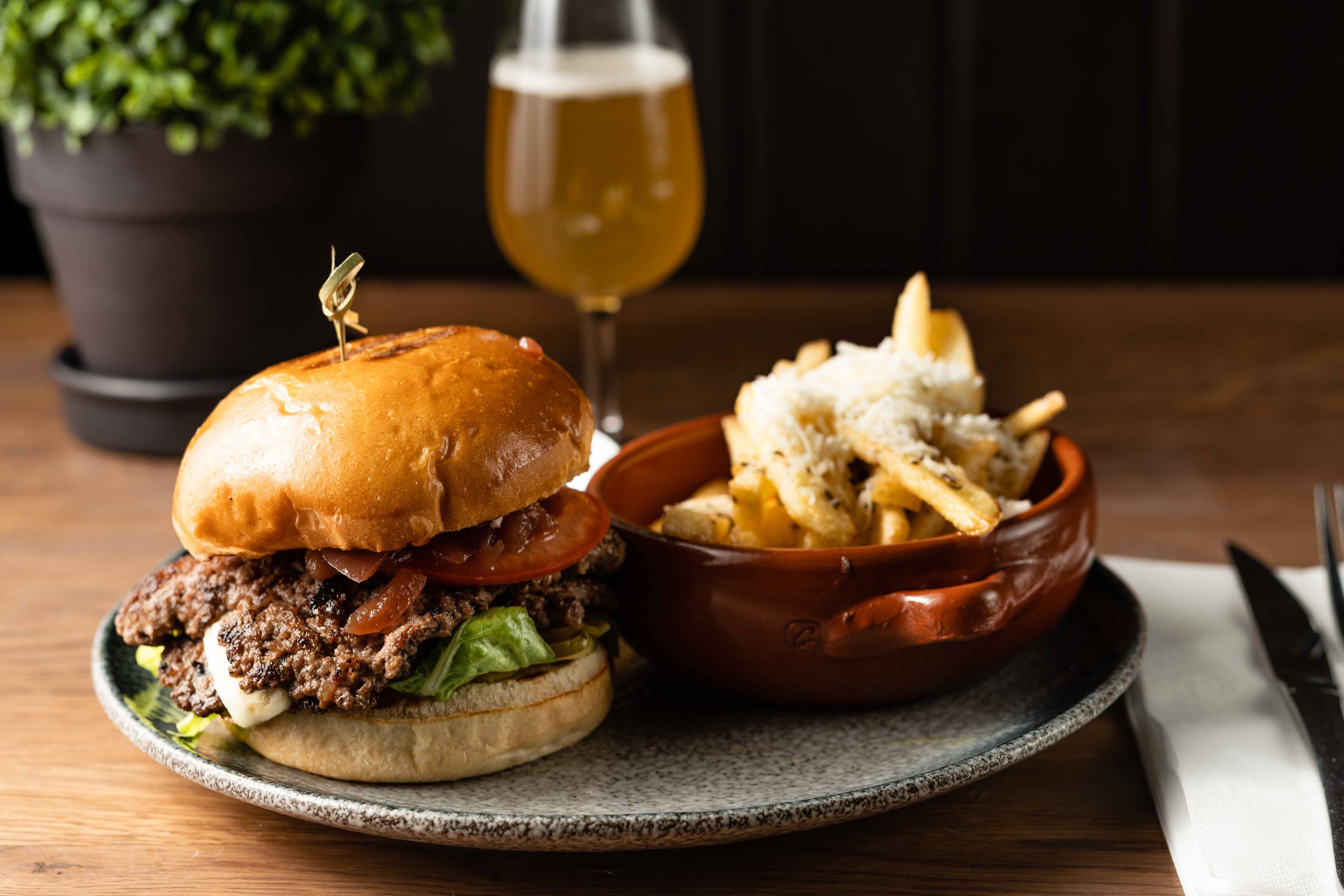 En saftig burger med ost, bacon og grønnsaker, servert med parmesanfries og et glass øl på en rustikk tallerken.