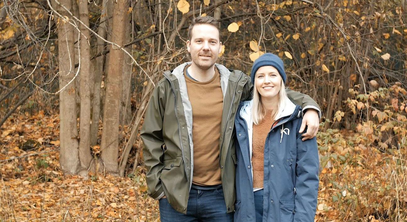 Et par som holder rundt hverandre og står ute blant høstløv og bare trær