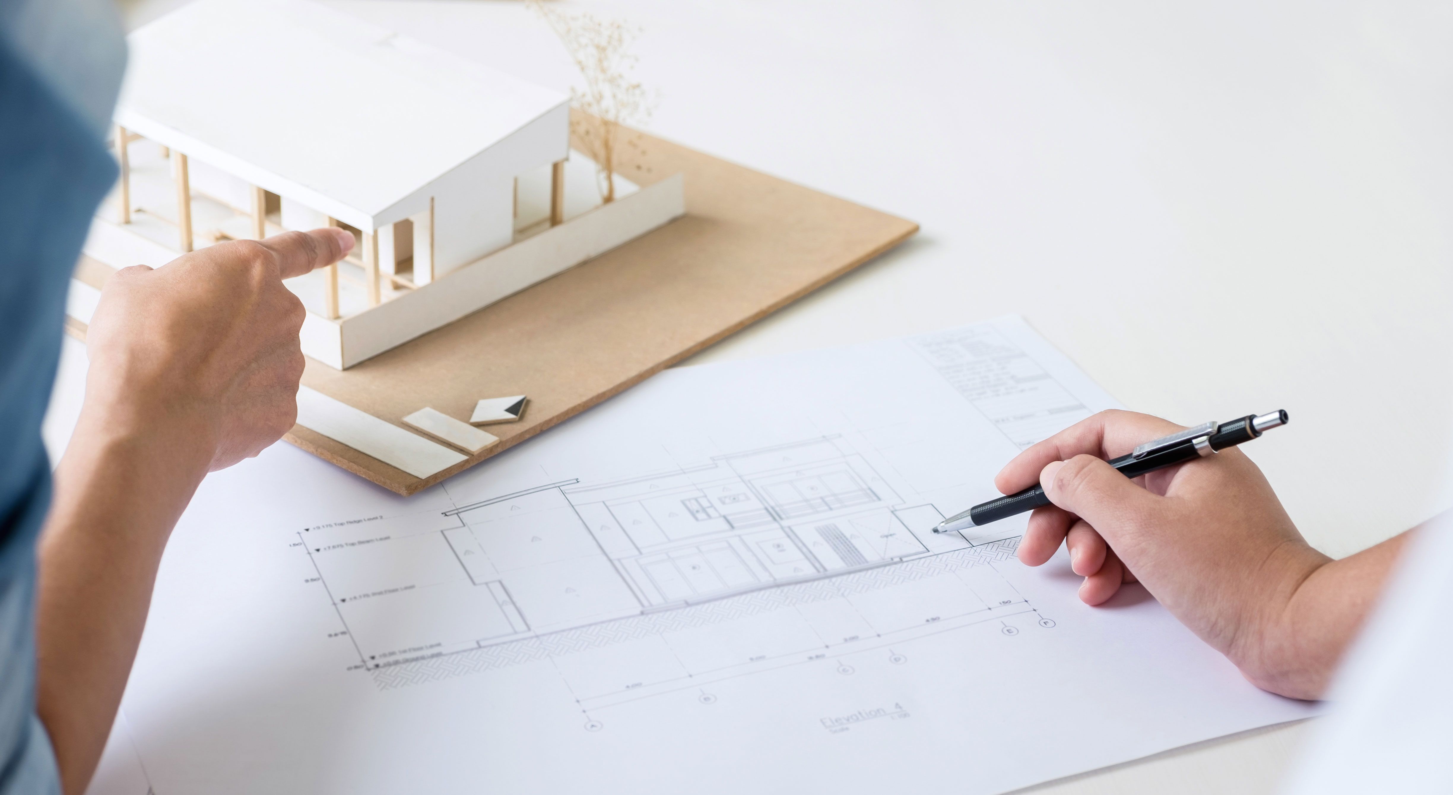 BIlde av to hender som tegner og peker på en arkitekttegning av et hus og en husmodell i papp