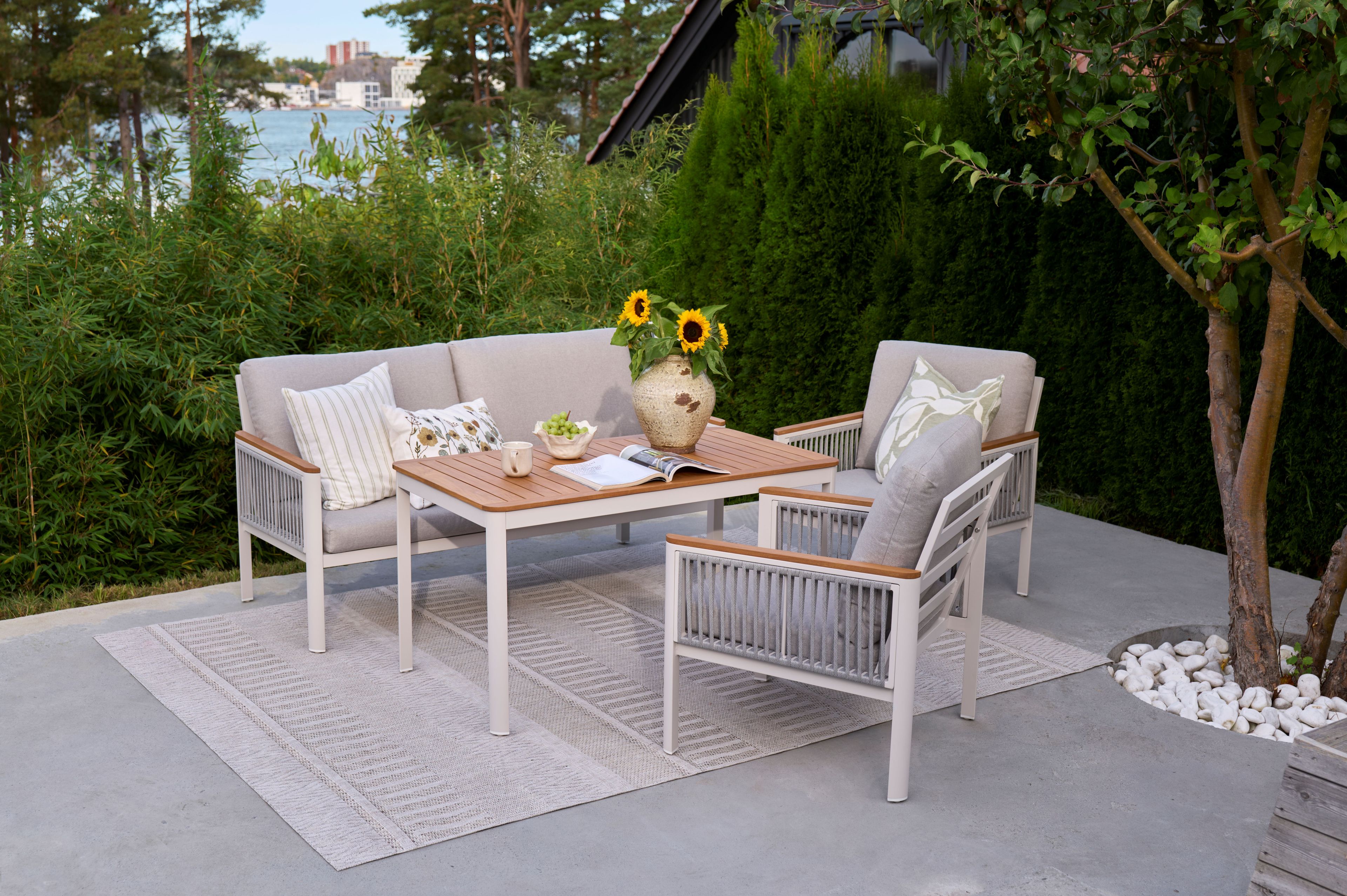 En sofa og to stoler står på en terrasse, på et teppe. Ved siden av er et tre og busker. Det står en vase på bordet med solsikker. Du skimter vannet fra sittegruppen.
