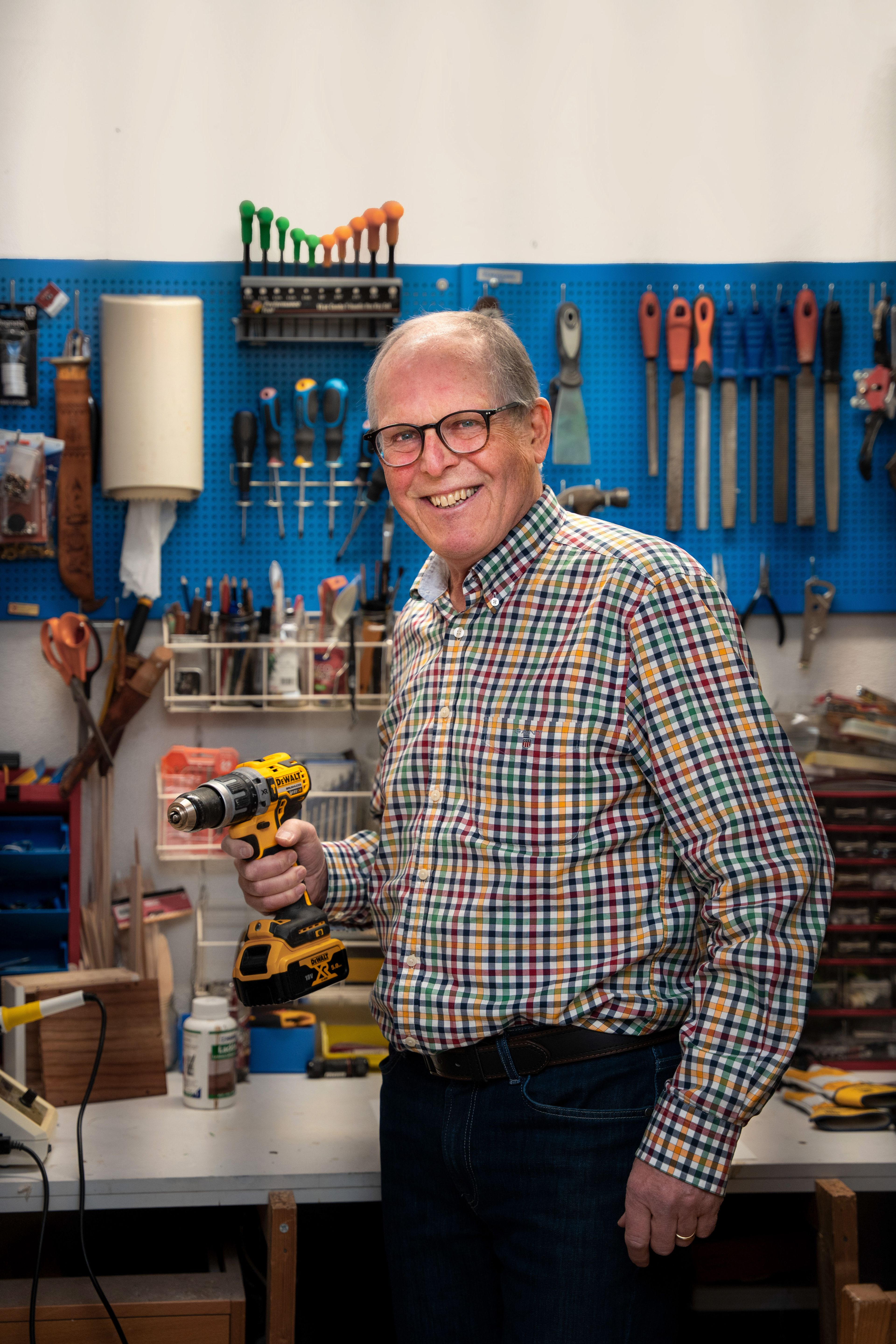 Mann i rutete skjorte står i verkstedet og holder en drill foran en vegg med verktøy.