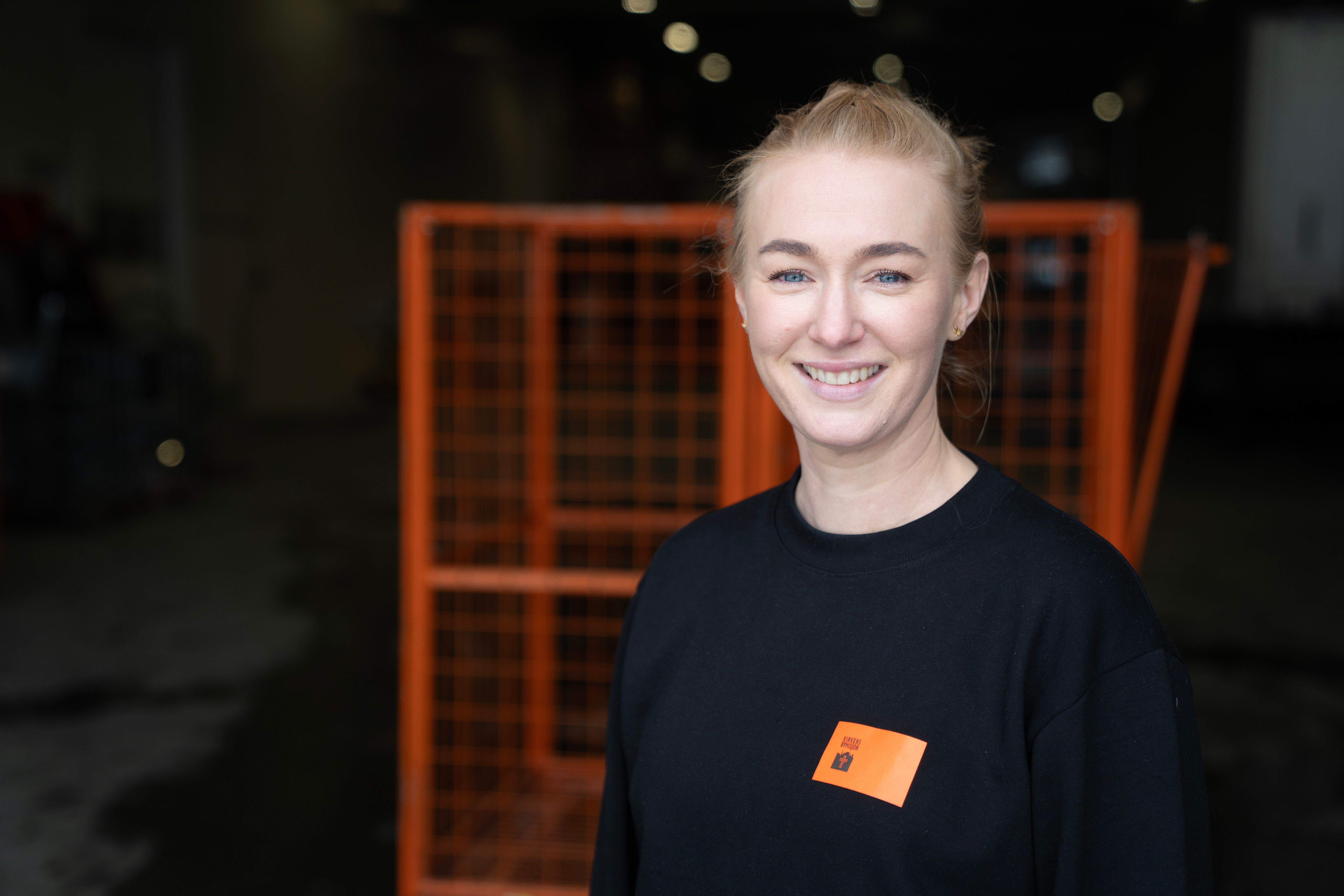 Kvinne som jobber hos Kirkens Bymisjon smiler foran oransje innsamlingsbur for klær.