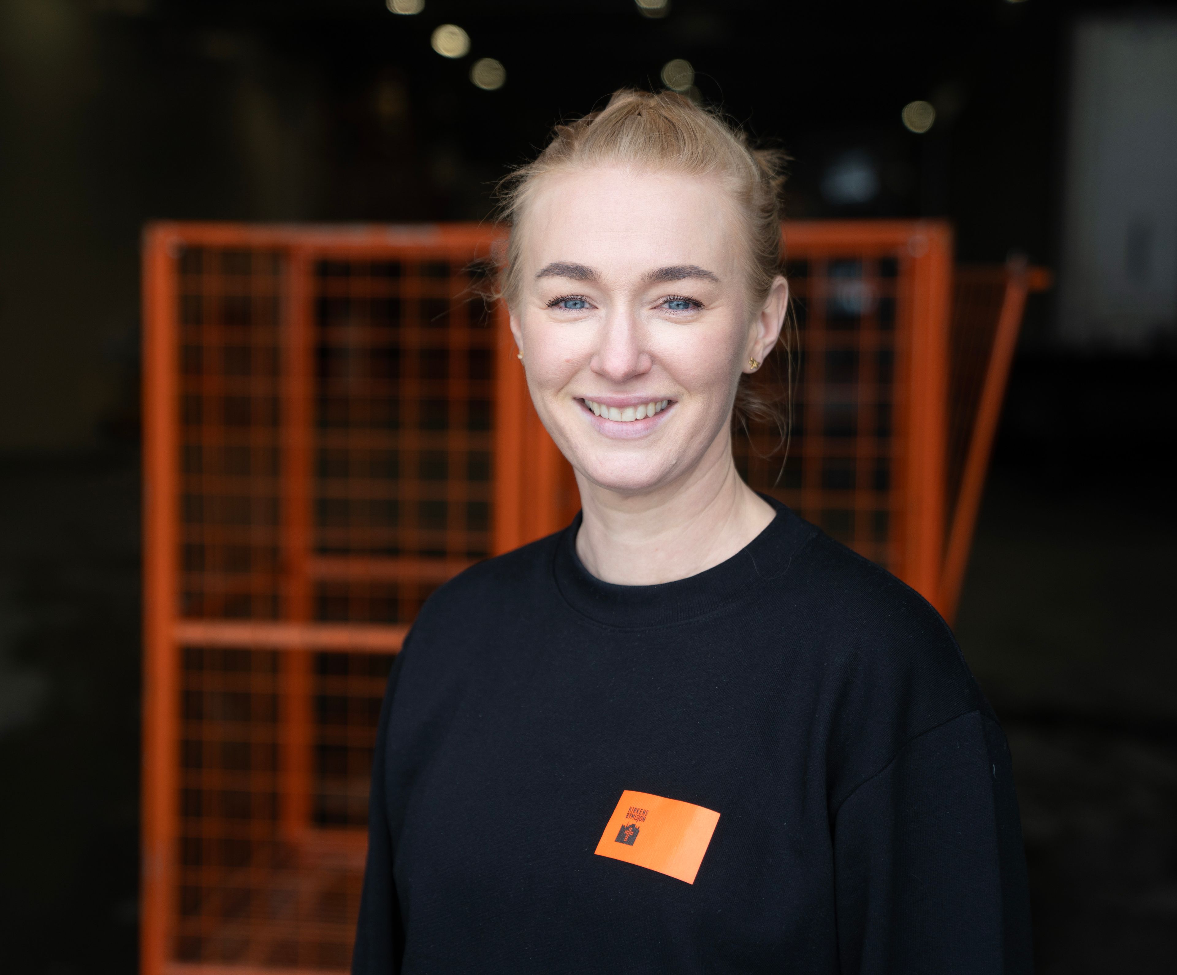 Kvinne som jobber hos Kirkens Bymisjon smiler foran oransje innsamlingsbur for klær.