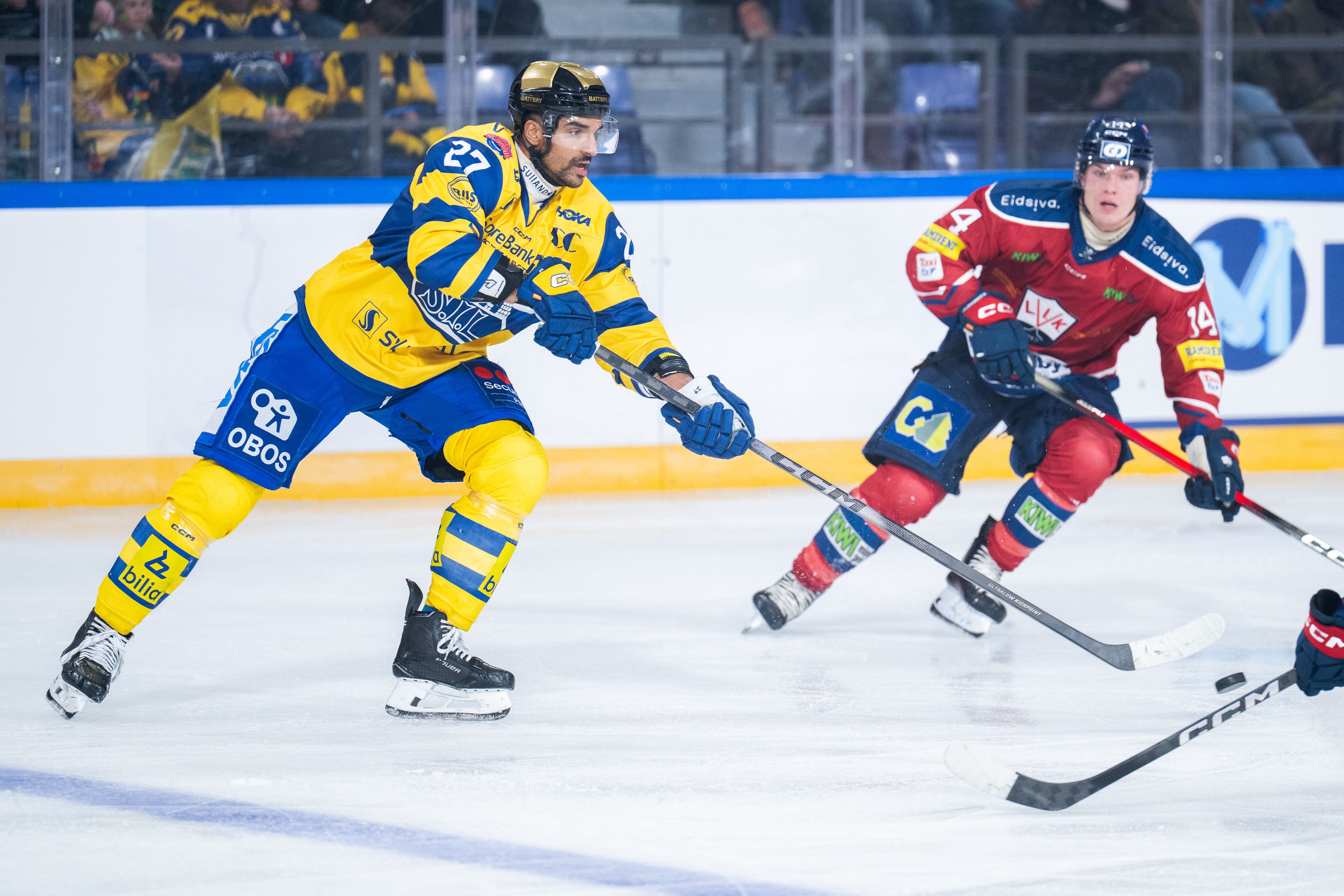 Ishockeyspiller i aksjon på banen i gul og blå drakt. I forgrunnen er hockeykøllen til en motspiller i rød og blå drakt, og bakgrunnen er en motspiller. 