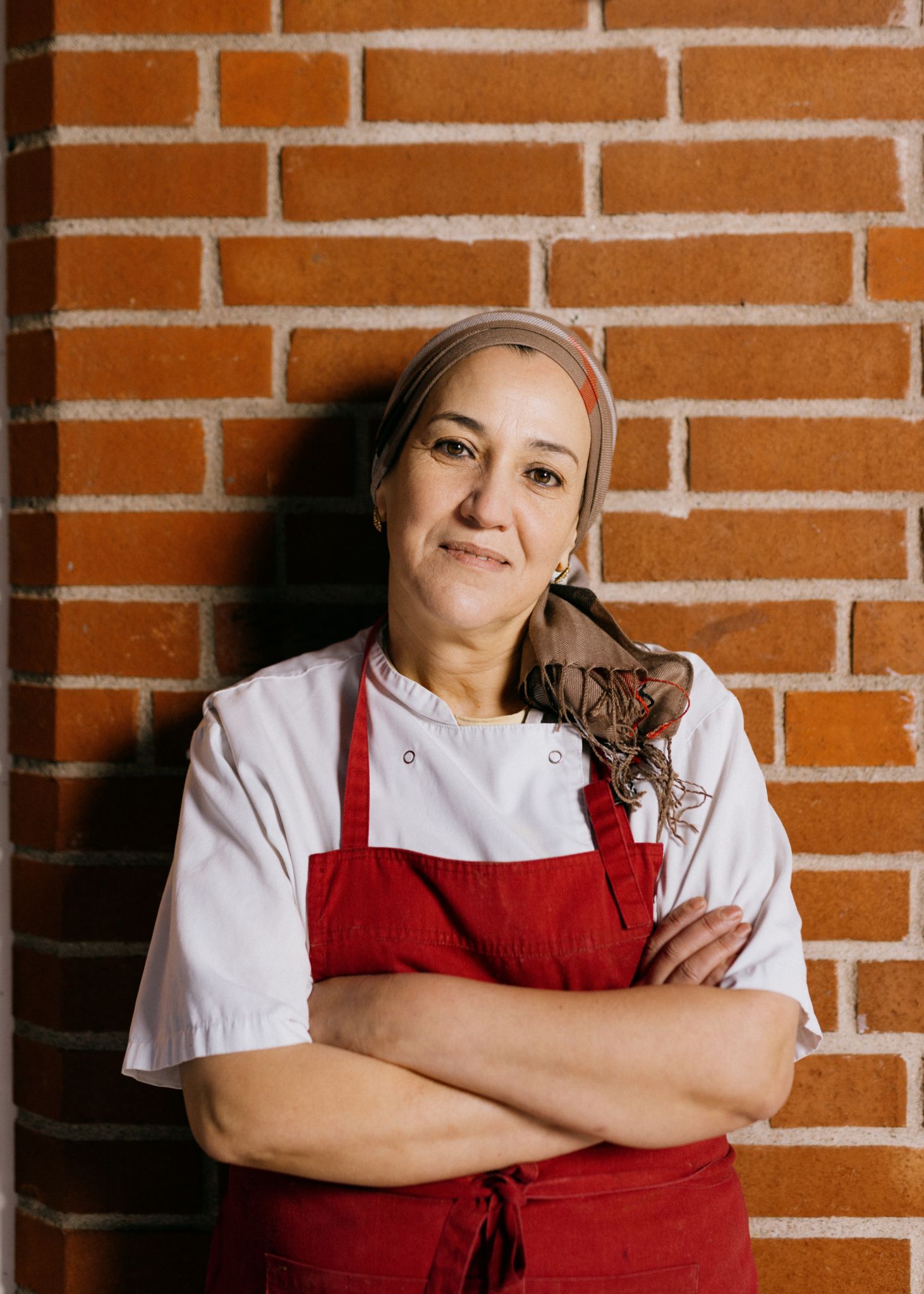 Foto av Nayat Abbouz som lener seg mot en rød murvegg mens hun har armene i kort og ser mot kameraet