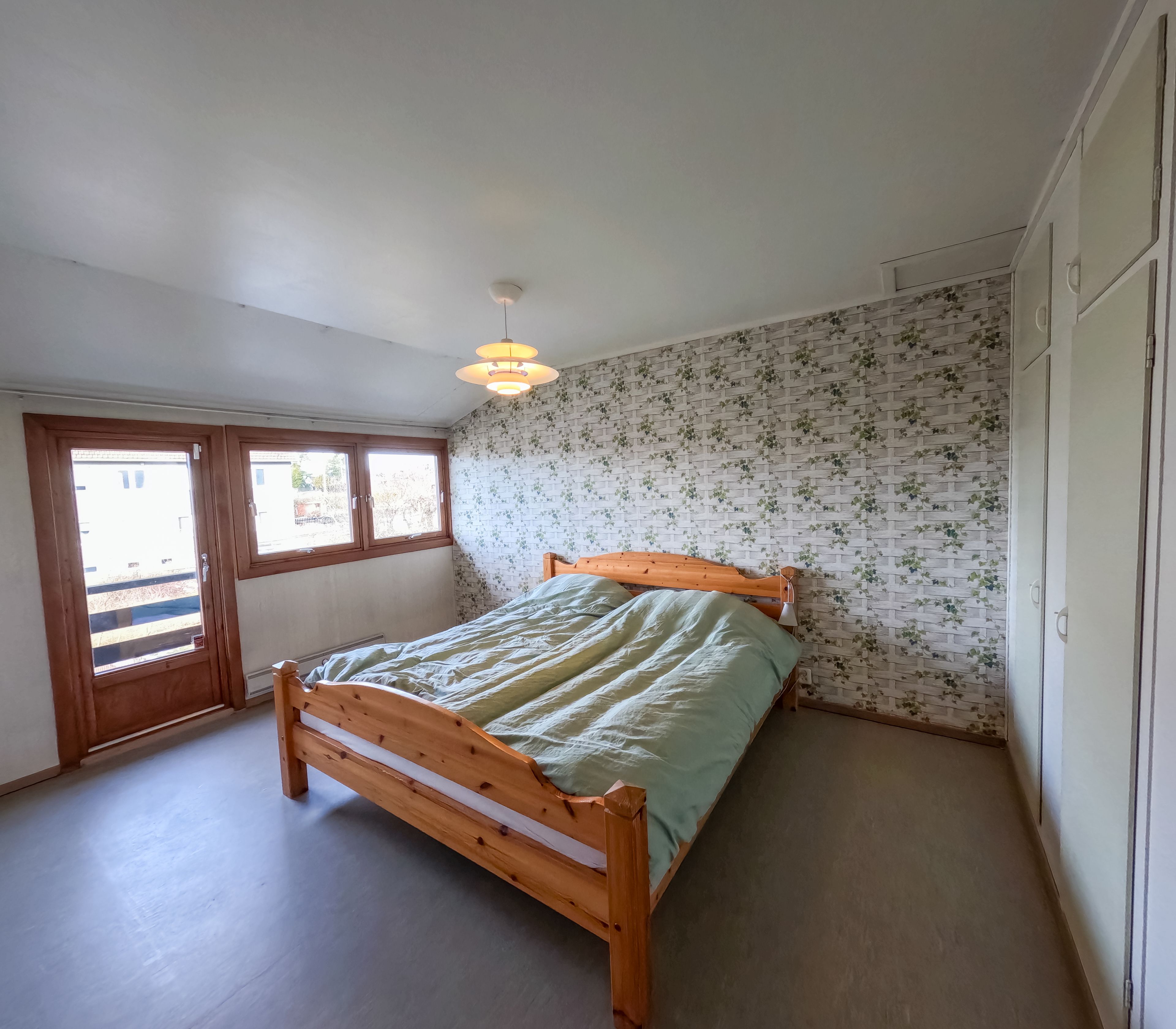 Eldre soverom med blomstrete tapet, linoleumsgulv og furuseng.