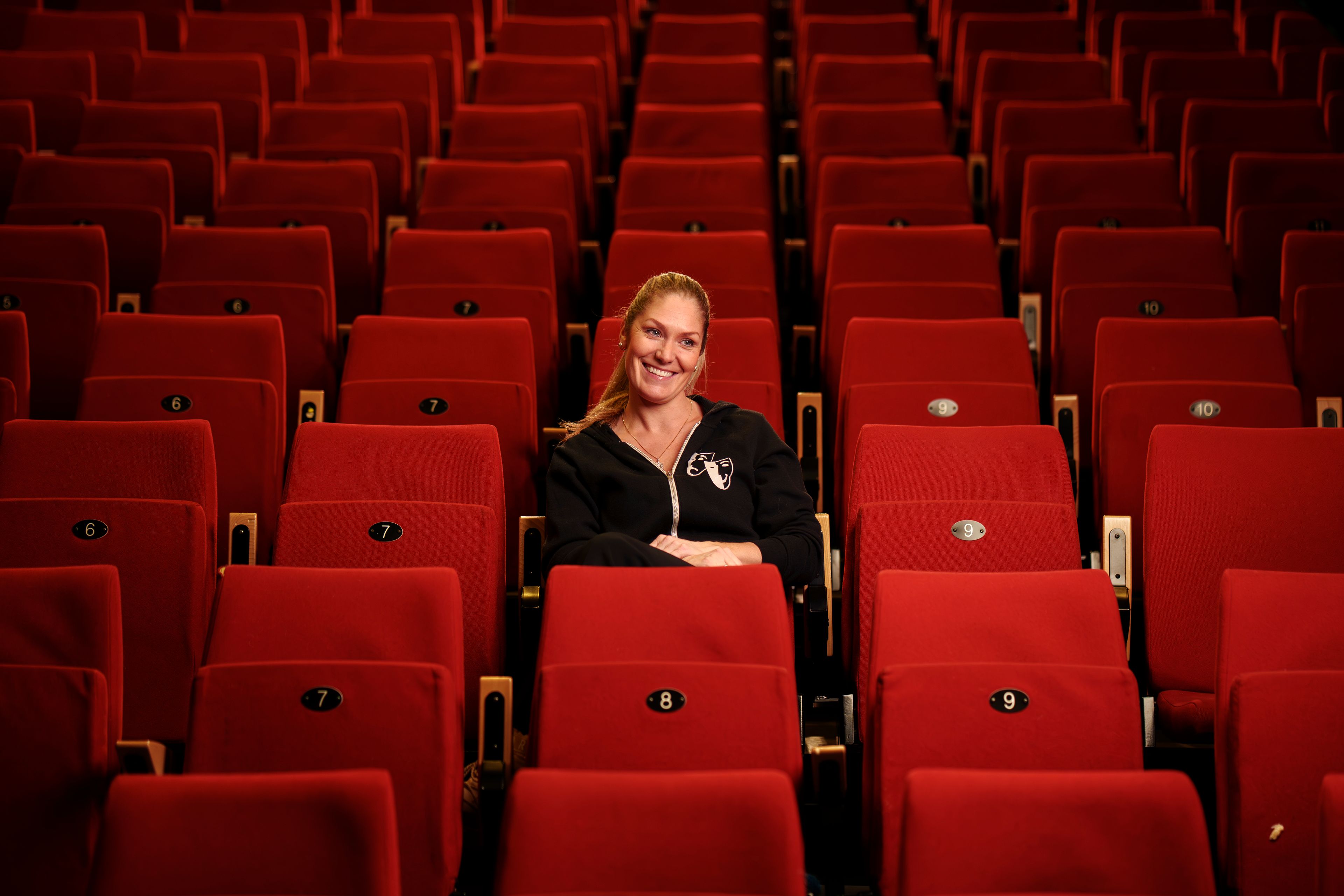 Mie Andberg sitter alene i en rad røde teaterstoler i salongen til Action Teater og smiler mot kameraet.