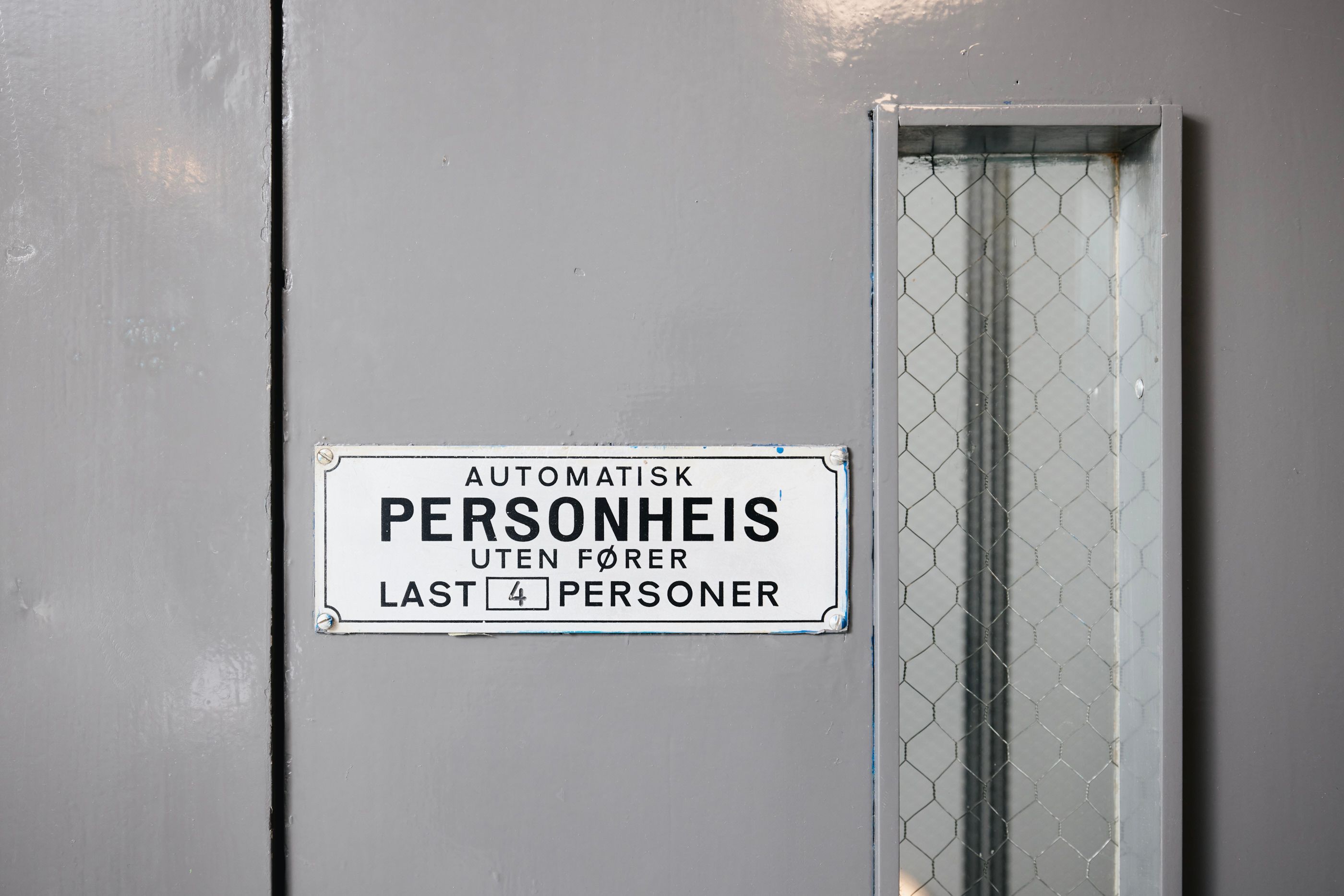 Heisdør i en boligblokk med skilt som viser at heisen er en automatisk personheis med kapasitet på fire personer.