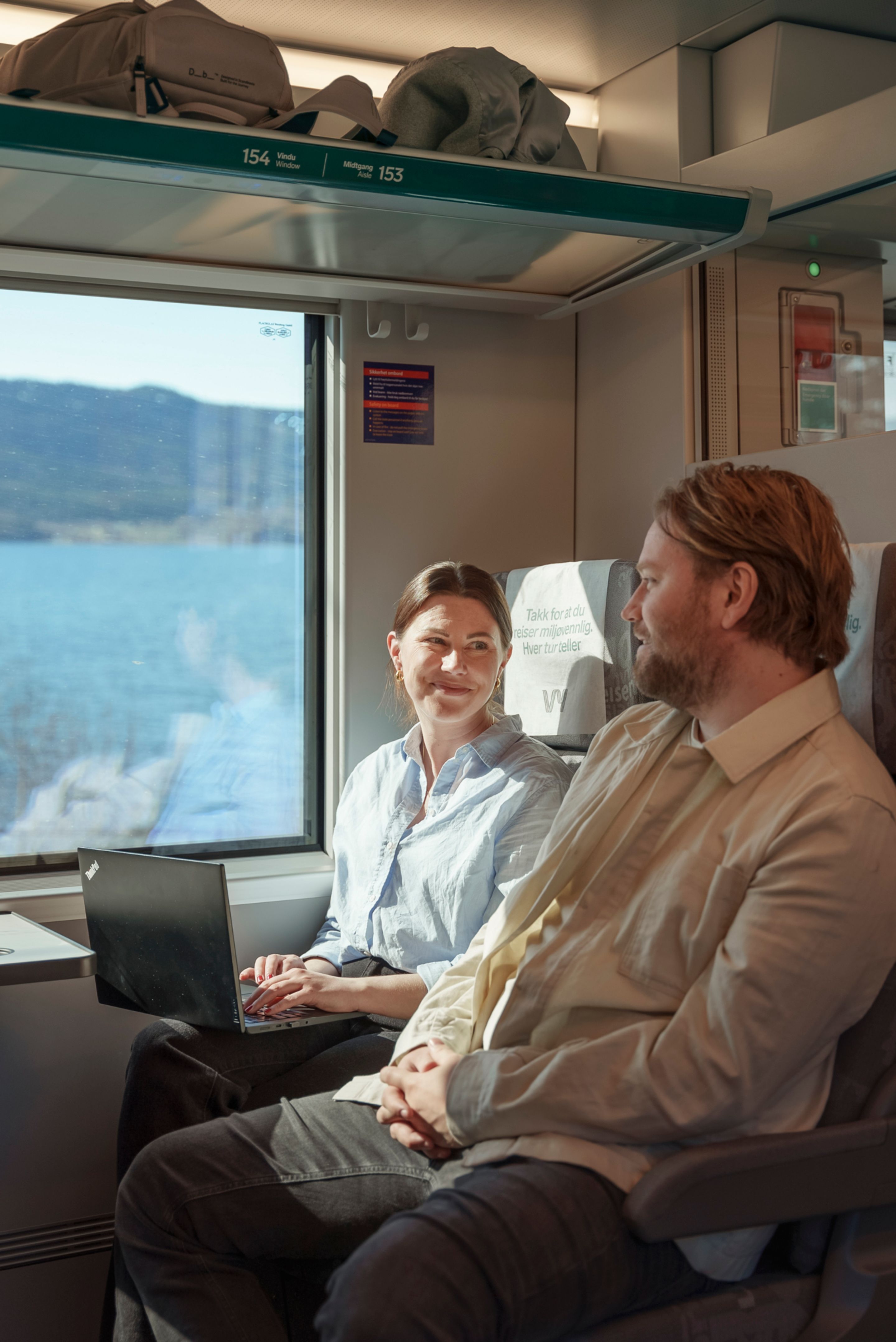 Stine og Martin snakker sammen på toget, med utsikt over Mjøsa i bakgrunnen.