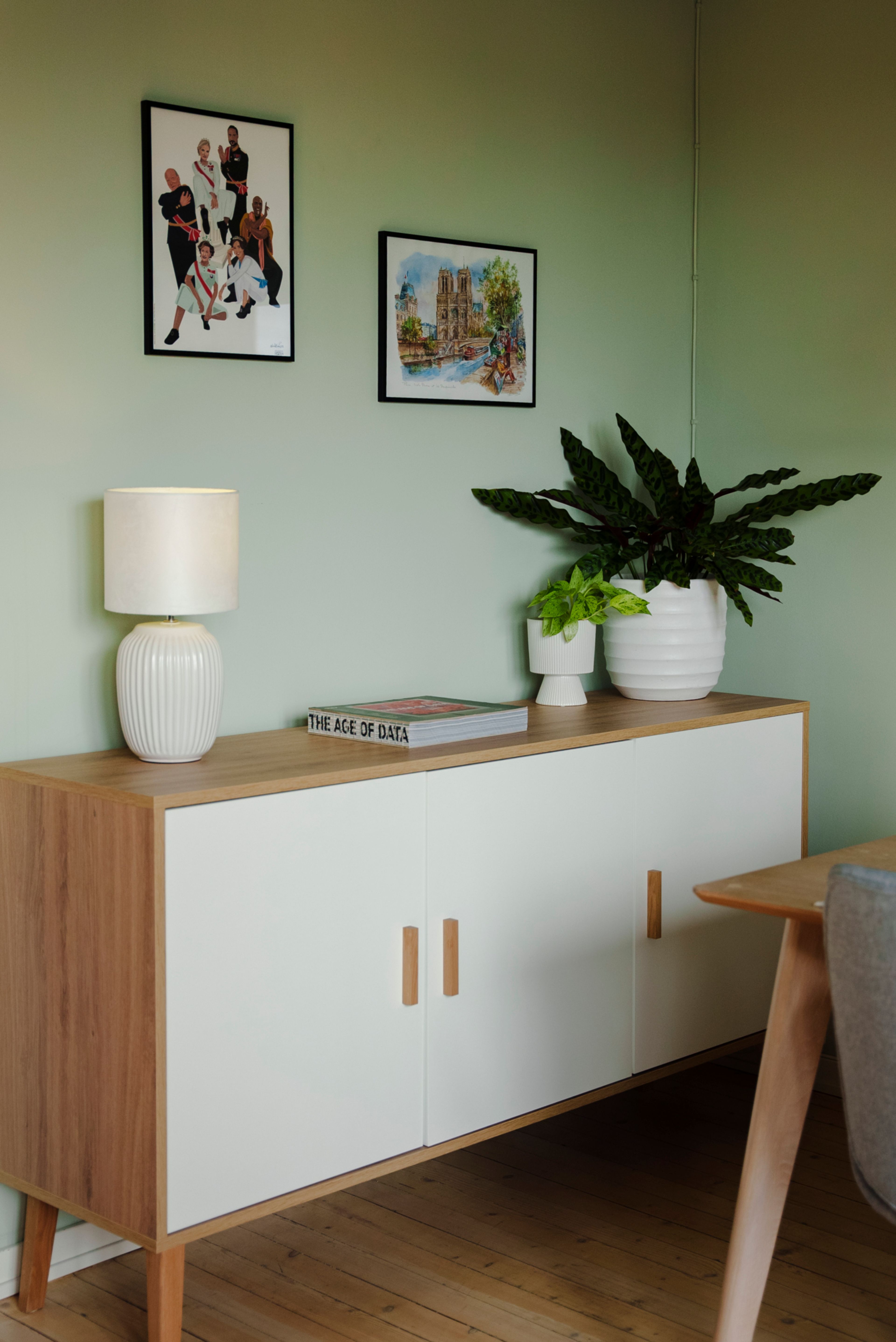 En hvit og trefarget skjenk i hjørnet av en stue, mot en grønnmalt vegg og noen planter og bilder.