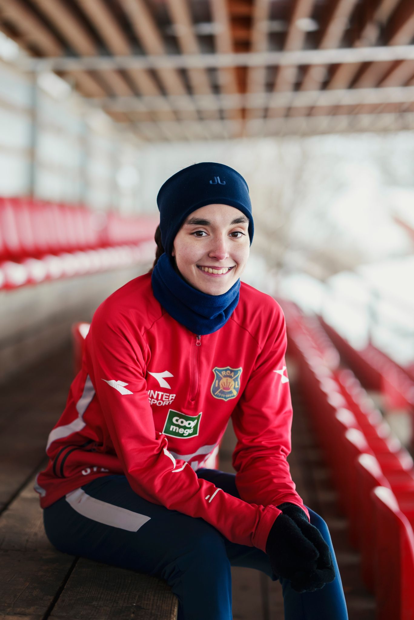 Sara sitter på de røde stolene på tribunen, hun smiler mot kamera.