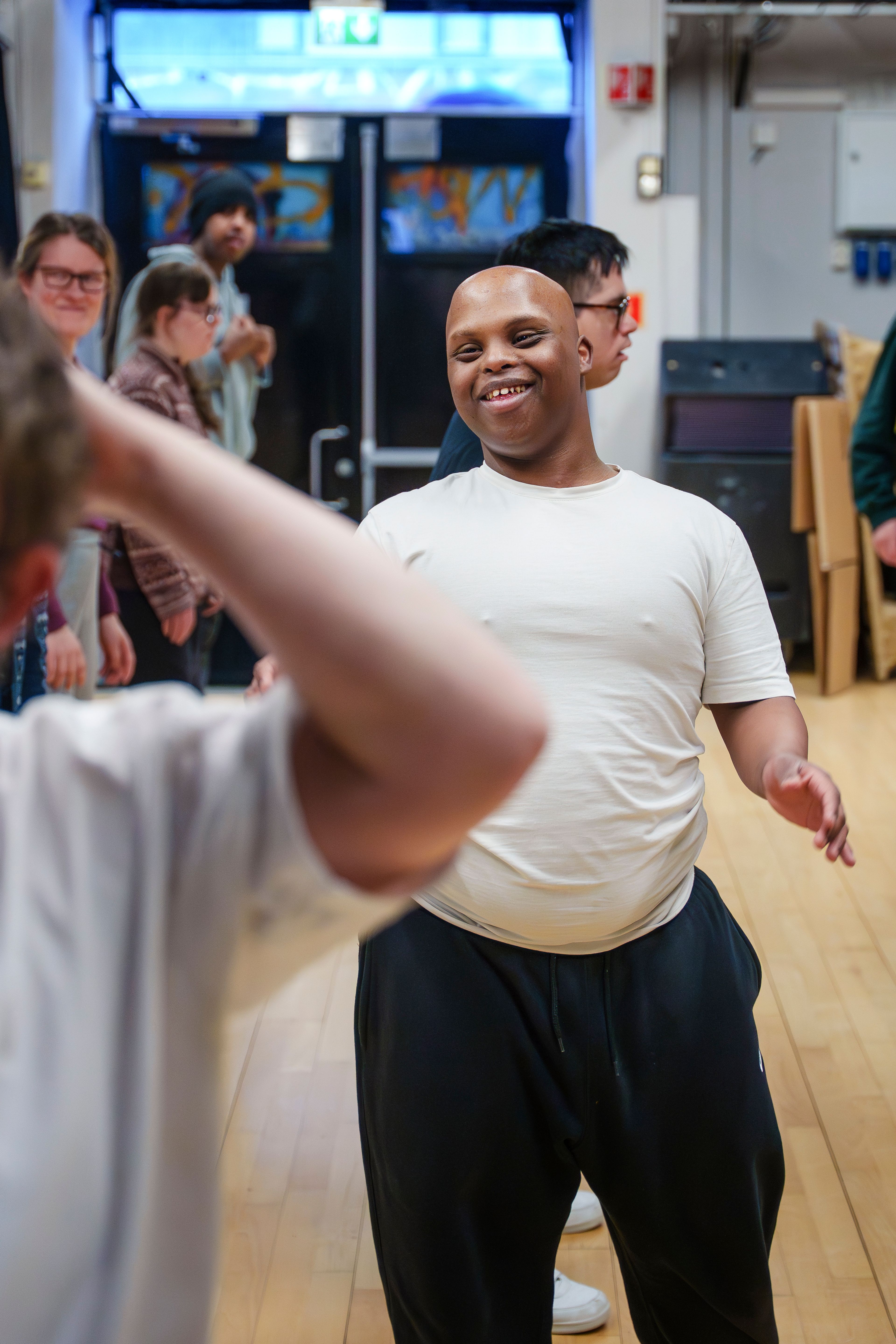 Smilende mann i hvit t-skjorte danser i et lyst rom mens en annen deltaker er i bevegelse i forgrunnen.