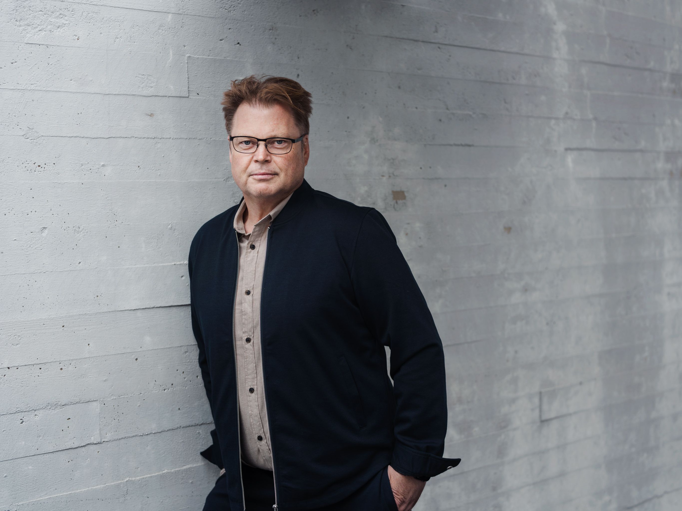 Foto av Jørn Lier Horst med en betongvegg i bakgrunn. Jørn har på seg en mørk beige skjorte, svarte jakke og briller.