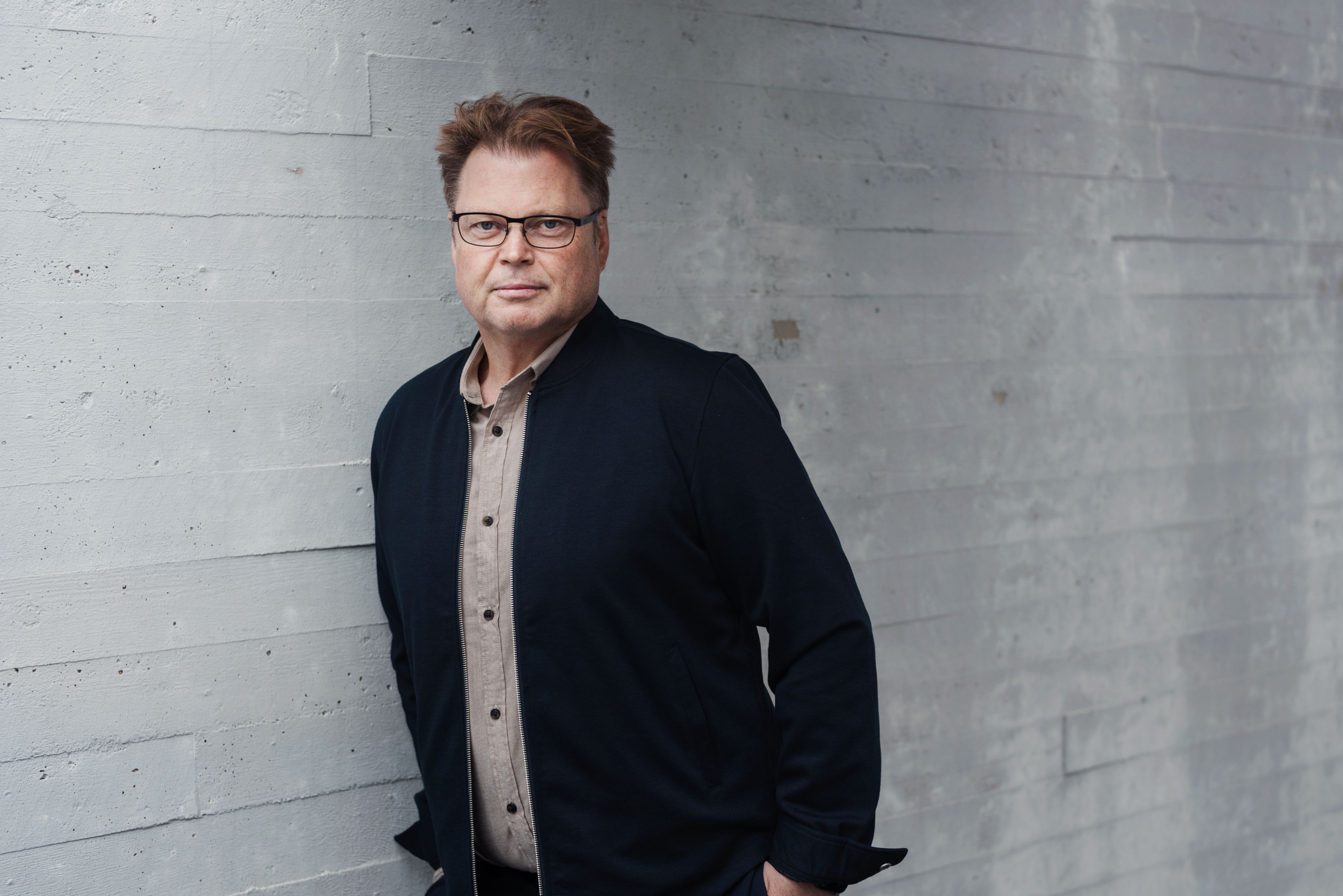Foto av Jørn Lier Horst med en betongvegg i bakgrunn. Jørn har på seg en mørk beige skjorte, svarte jakke og briller.