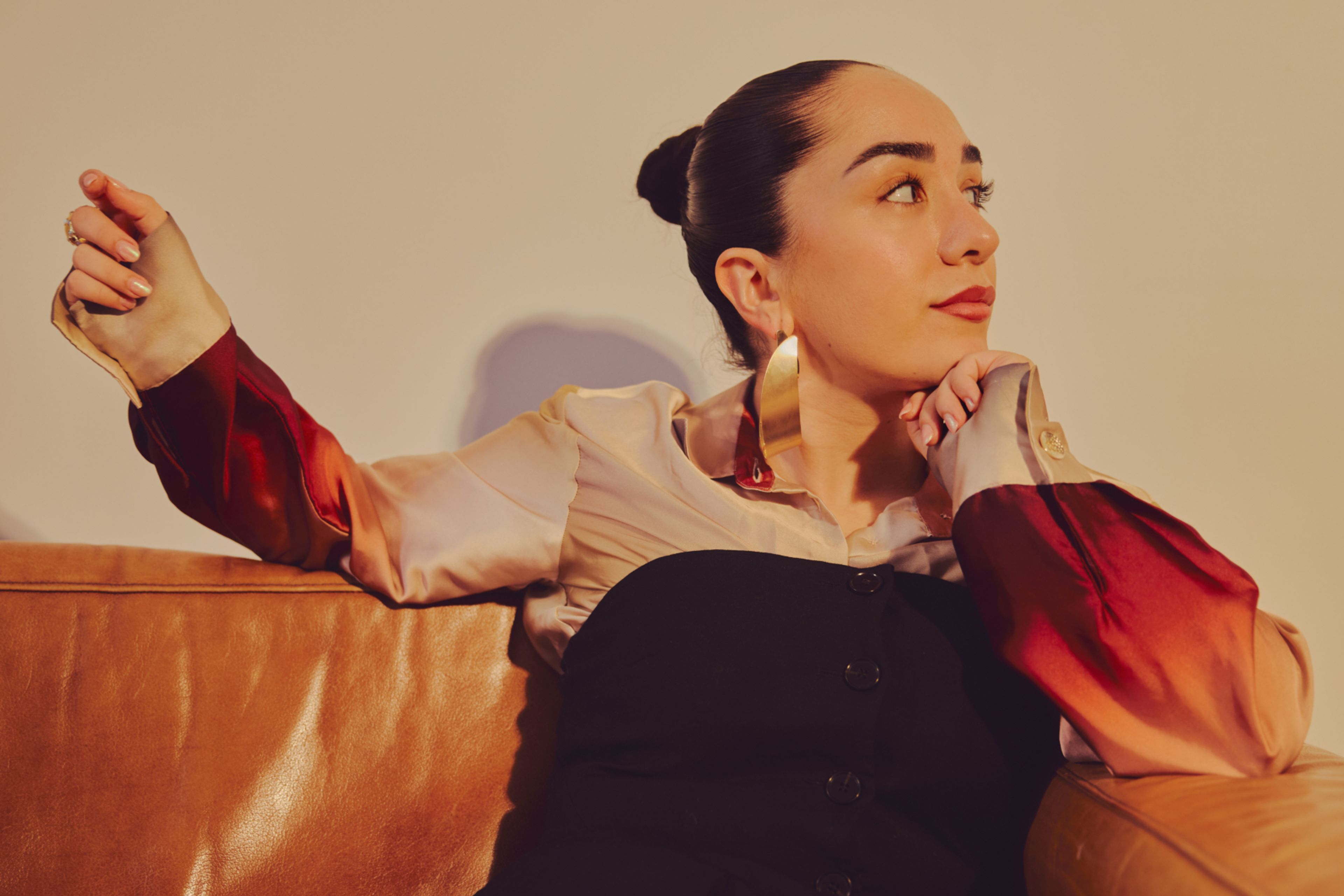 Amanda Delara sitter i en brun skinnsofa. Hun har på seg en silkeskjorte i gull, med en svart kjole over. Hun har store gulløredobber i ørene og håret satt opp i en stram, elegant knute.