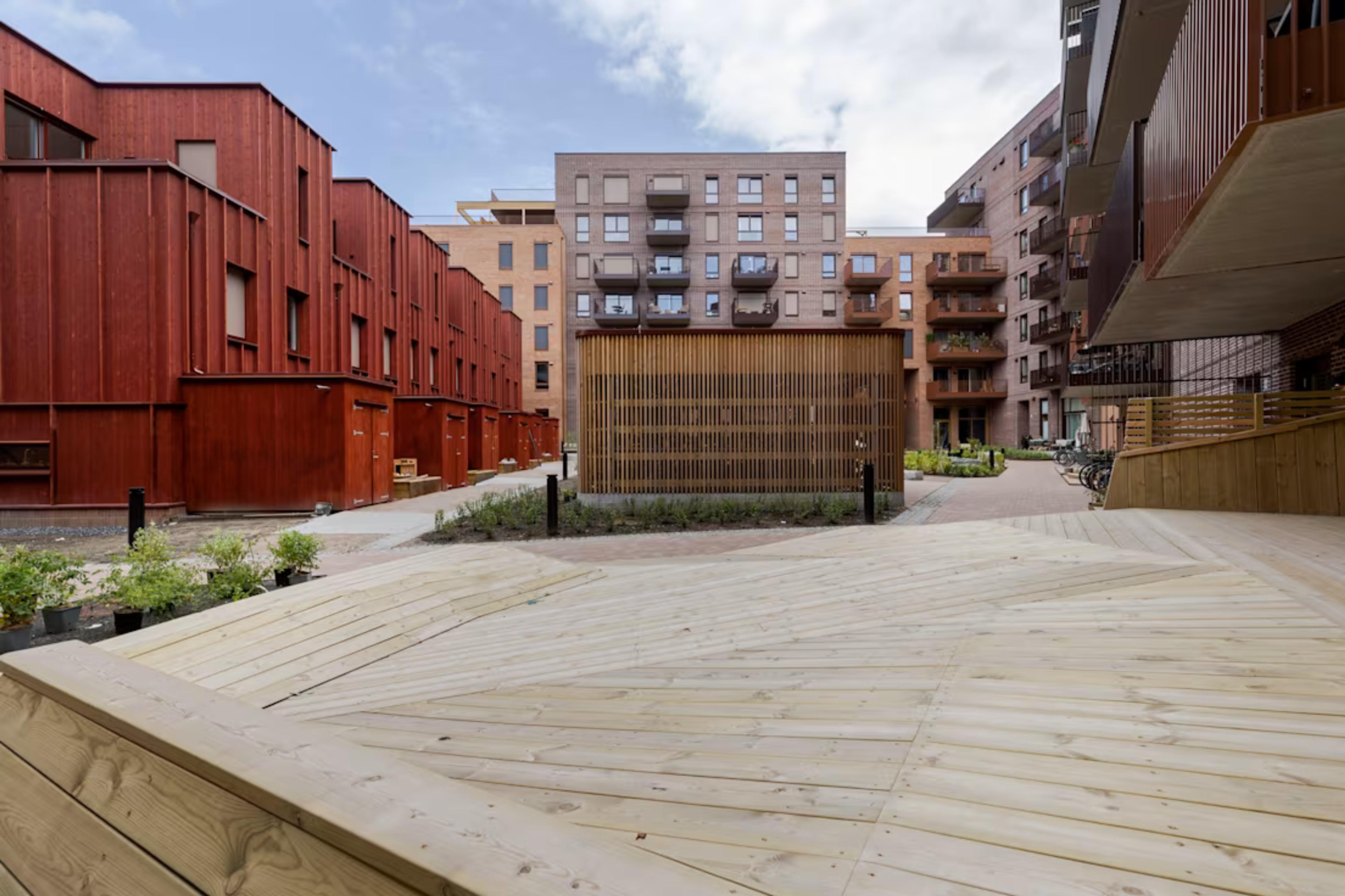 Moderne bygård med røde og brune bygninger rundt. Trestier og blomsterkasser skaper en innbydende og moderne atmosfære under en delvis overskyet himmel.