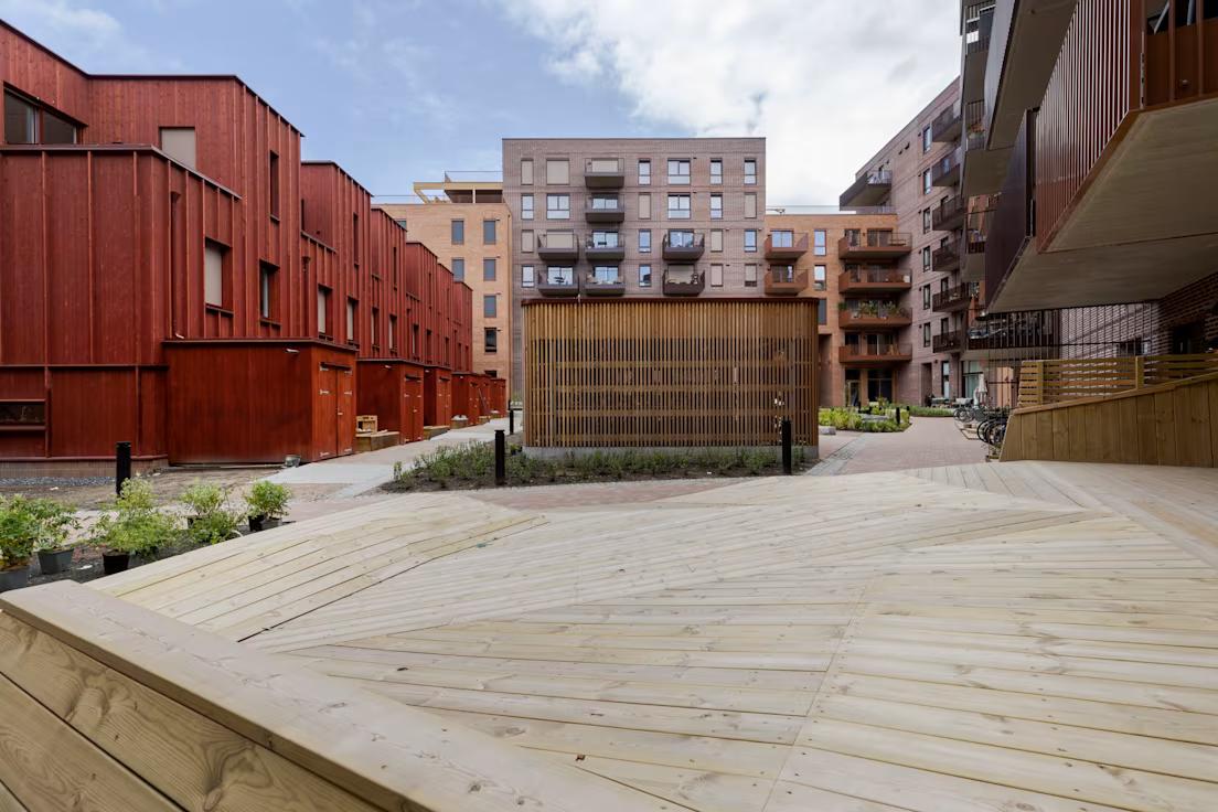 Moderne bygård med røde og brune bygninger rundt. Trestier og blomsterkasser skaper en innbydende og moderne atmosfære under en delvis overskyet himmel.