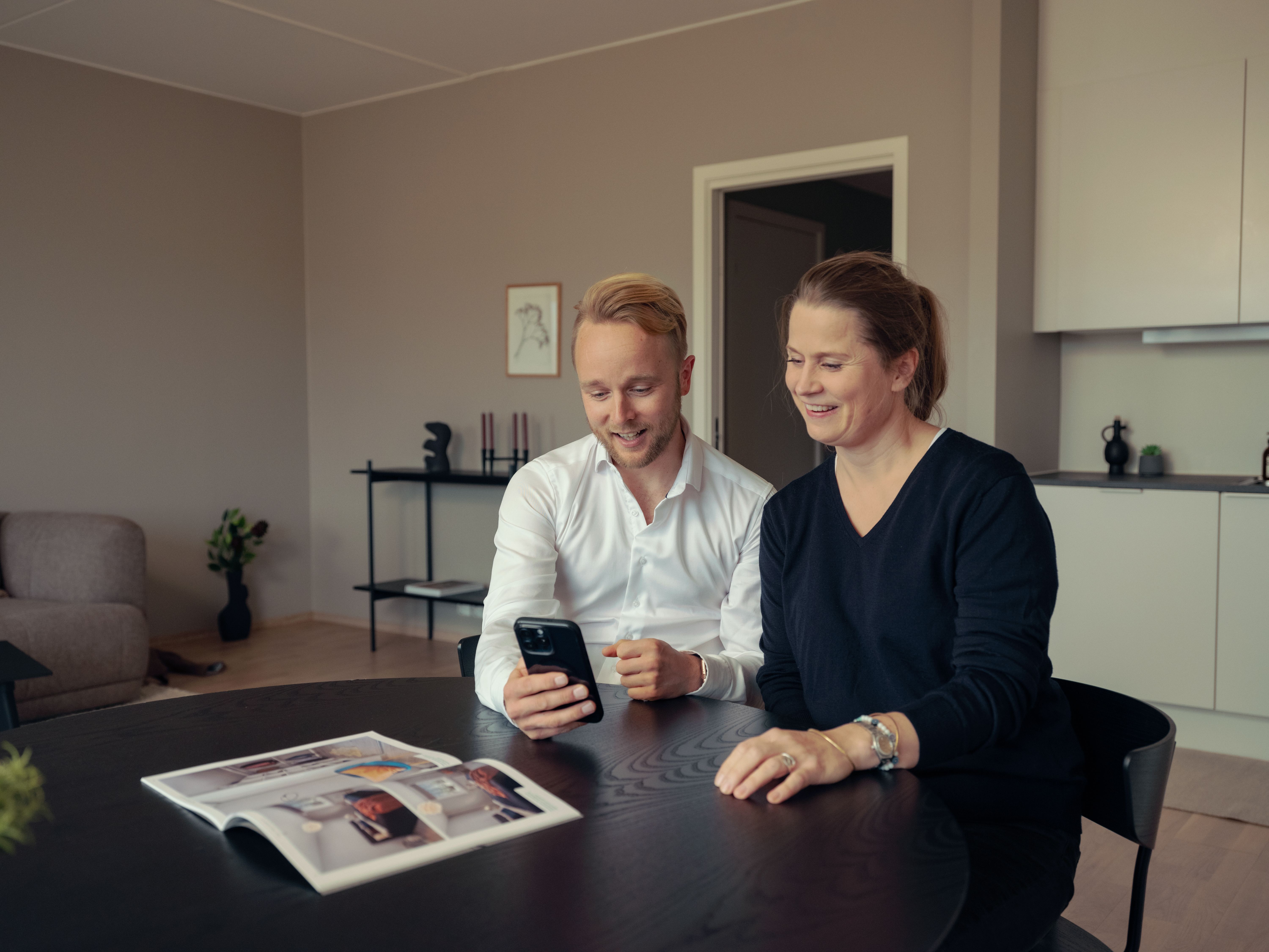To personer sitter ved et spisebord i en leilighet og ser på mobiltelefonen og smiler. På bordet ligger en brosjyre.