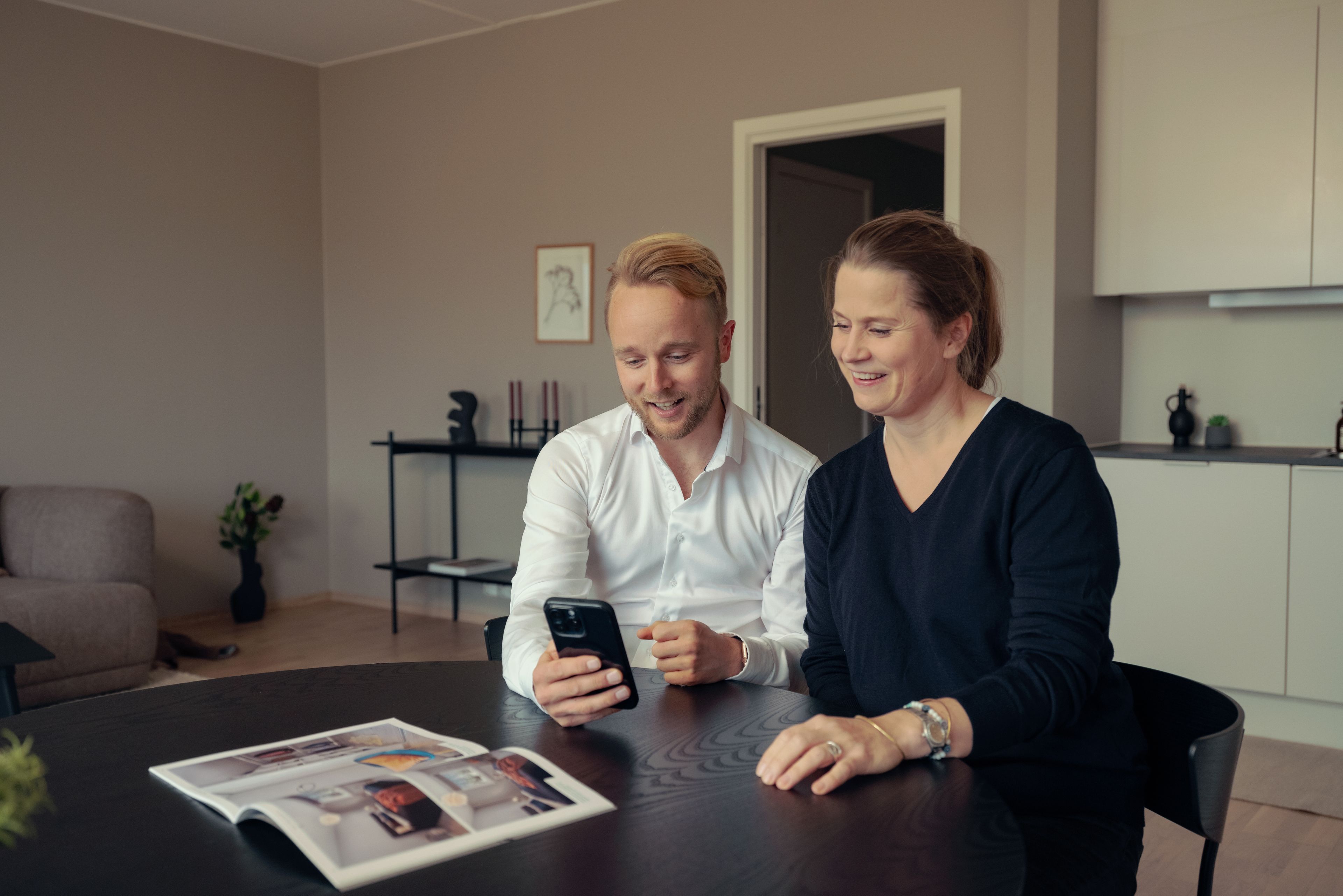 To personer sitter ved et spisebord i en leilighet og ser på mobiltelefonen og smiler. På bordet ligger en brosjyre.