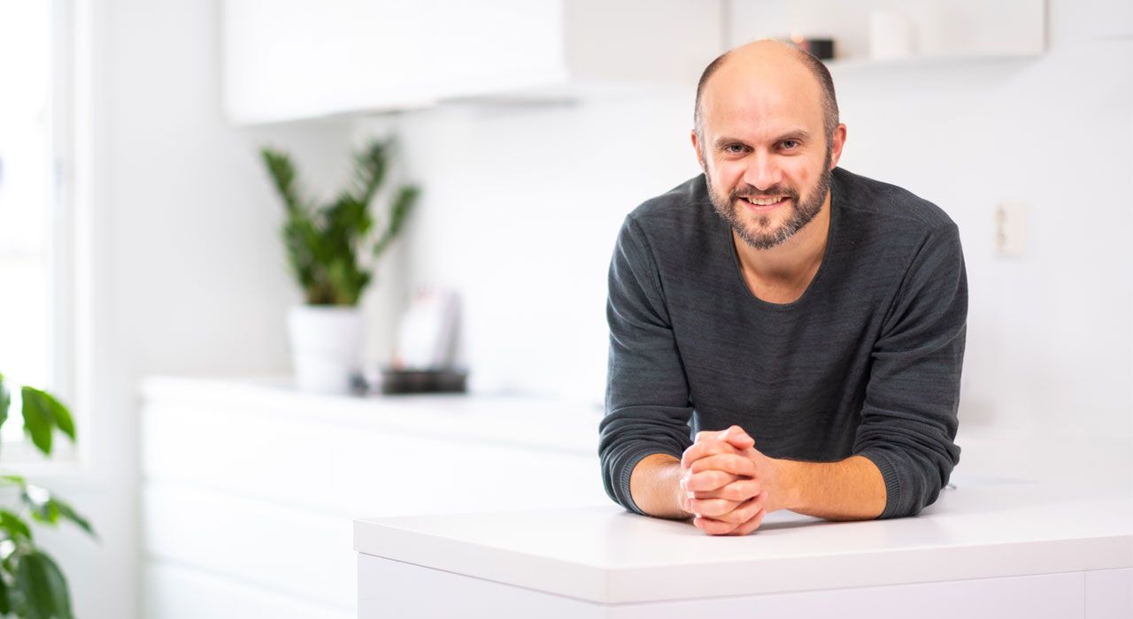 Fornøyd mann som lener seg smilende over en hvit kjøkkenbenk og ser inni kameraet.