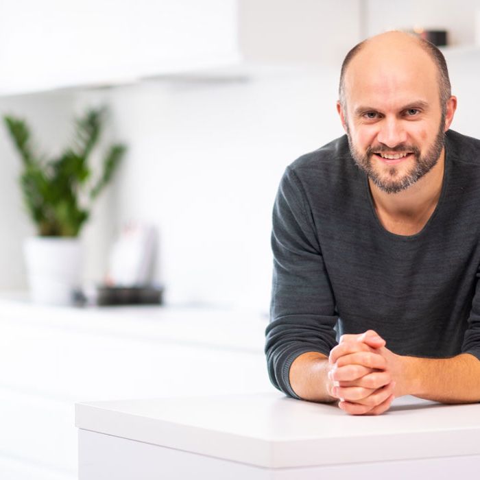 Fornøyd mann som lener seg smilende over en hvit kjøkkenbenk og ser inni kameraet.