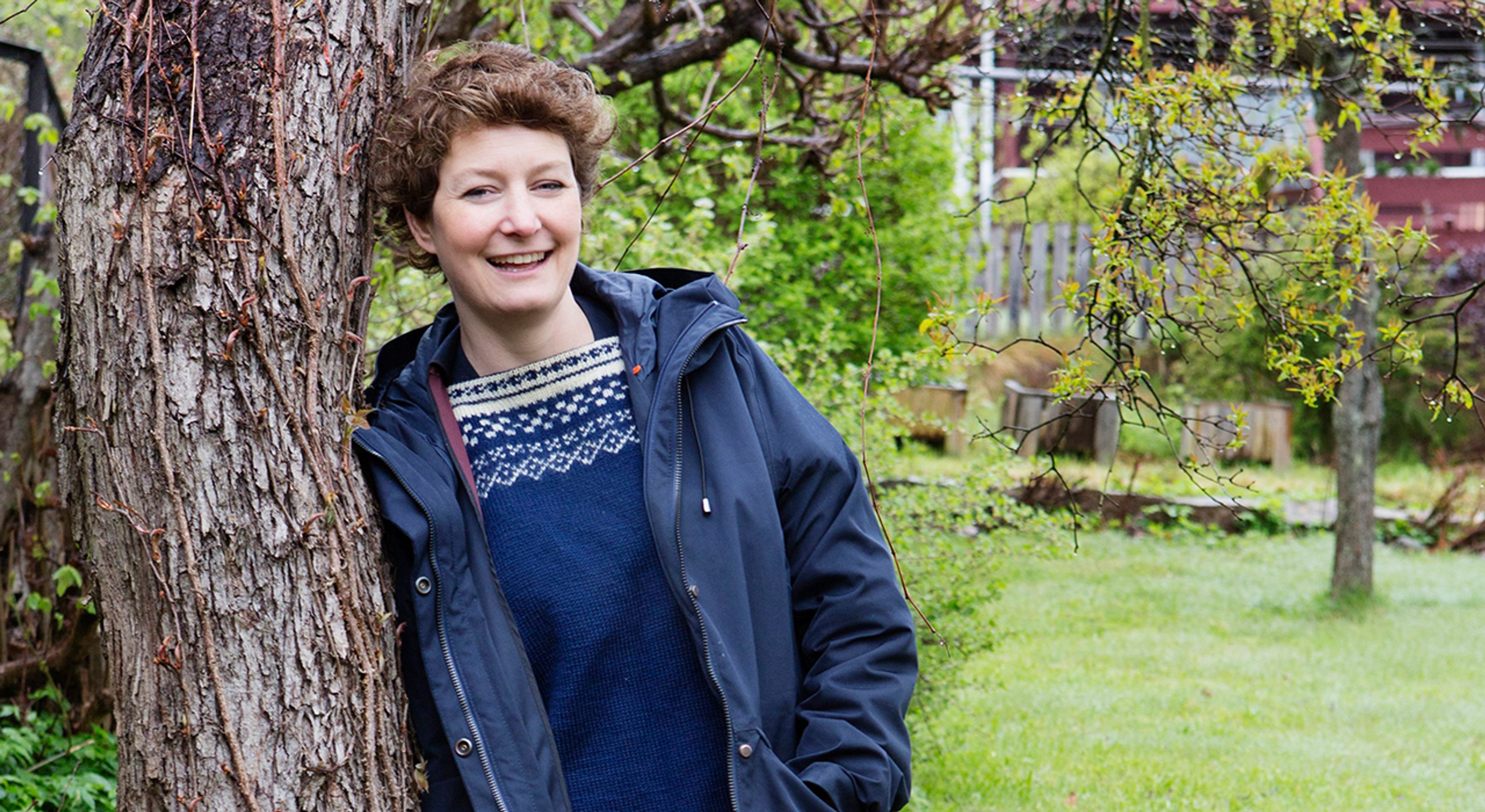 En kvinne med brunt hår lener seg inntil et tre og smiler til kamera.