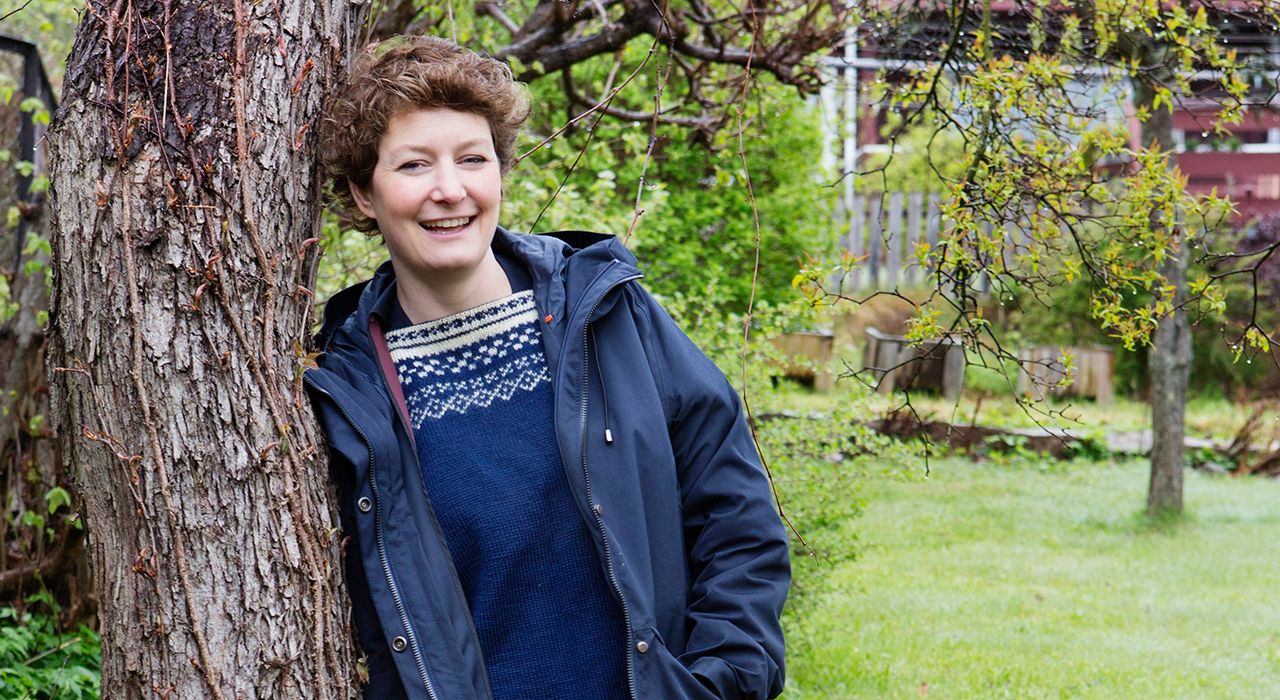 En kvinne med brunt hår lener seg inntil et tre og smiler til kamera.