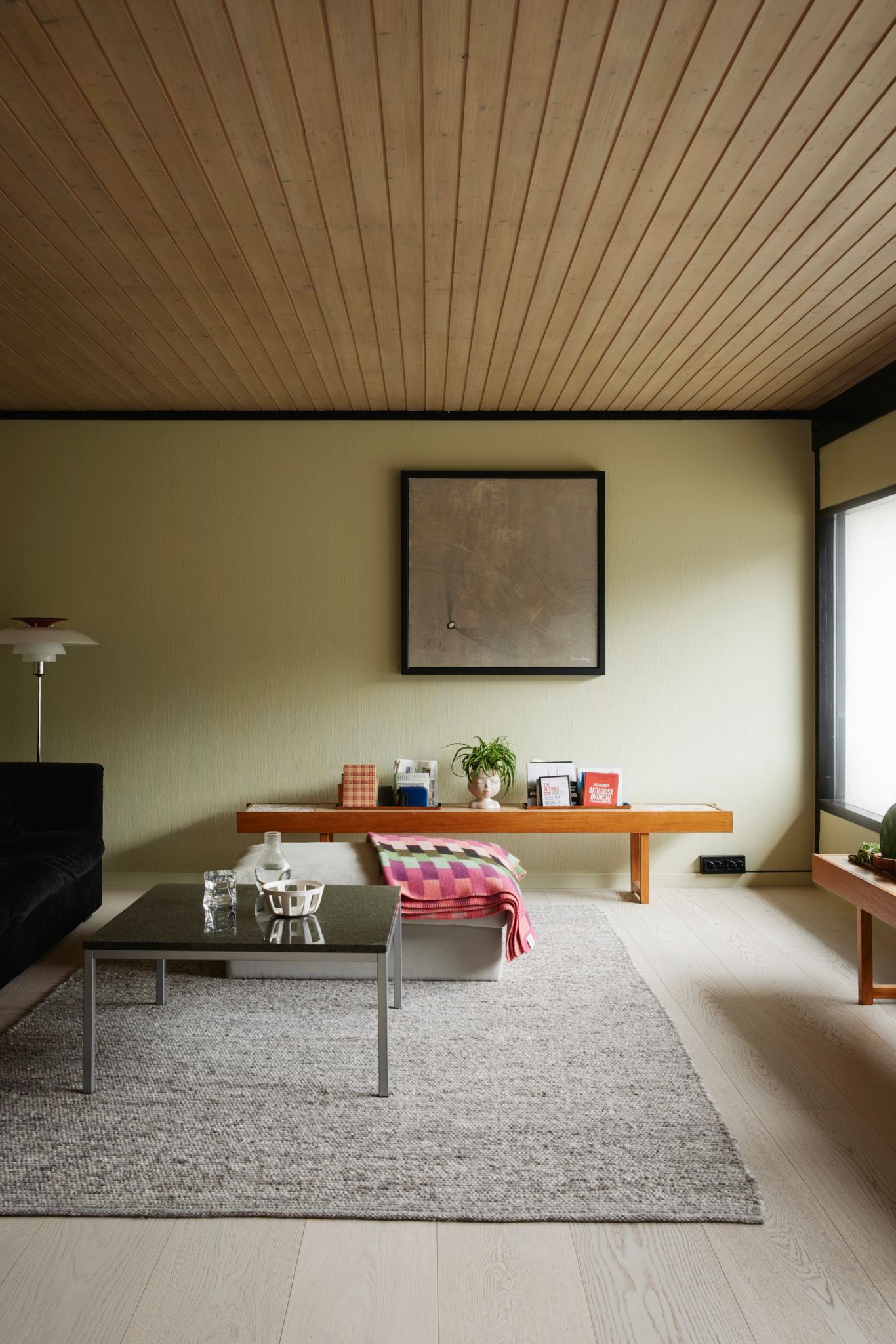 En stue med en minimalistisk innredning, inkludert et lavt sofabord på et grått teppe og en trebenk langs veggen med bøker og dekor. Veggene er lyse, og et kunstverk henger over benken.