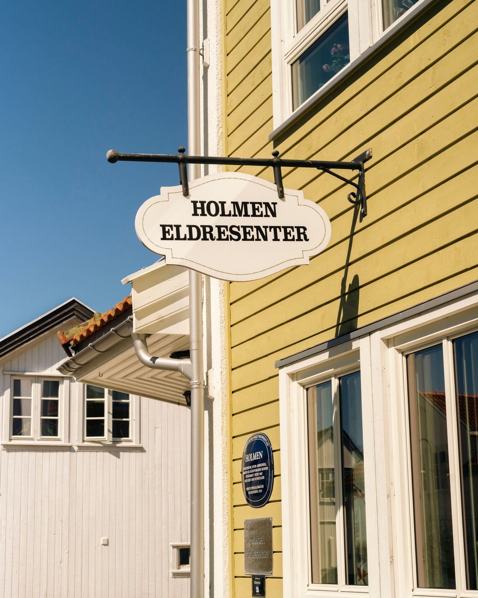 Hvitt skilt med teksten «Holmen Eldresenter» henger i smijernsfeste på gul trevegg mot blå himmel.