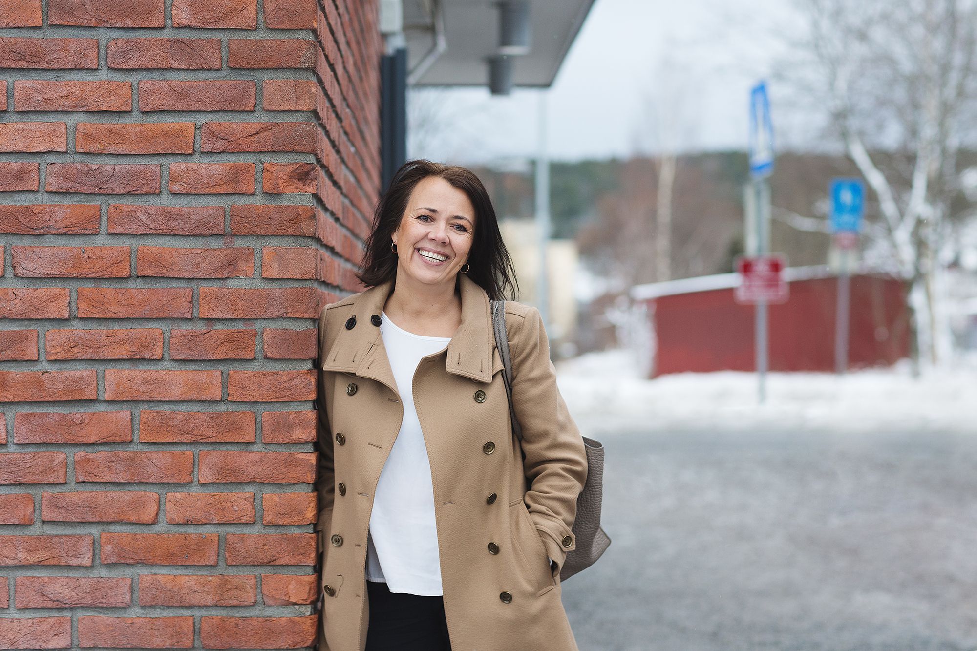 Eiendomsmegler Mona Pedersen smiler til kameraet mens hun lener seg mot en rød murvegg