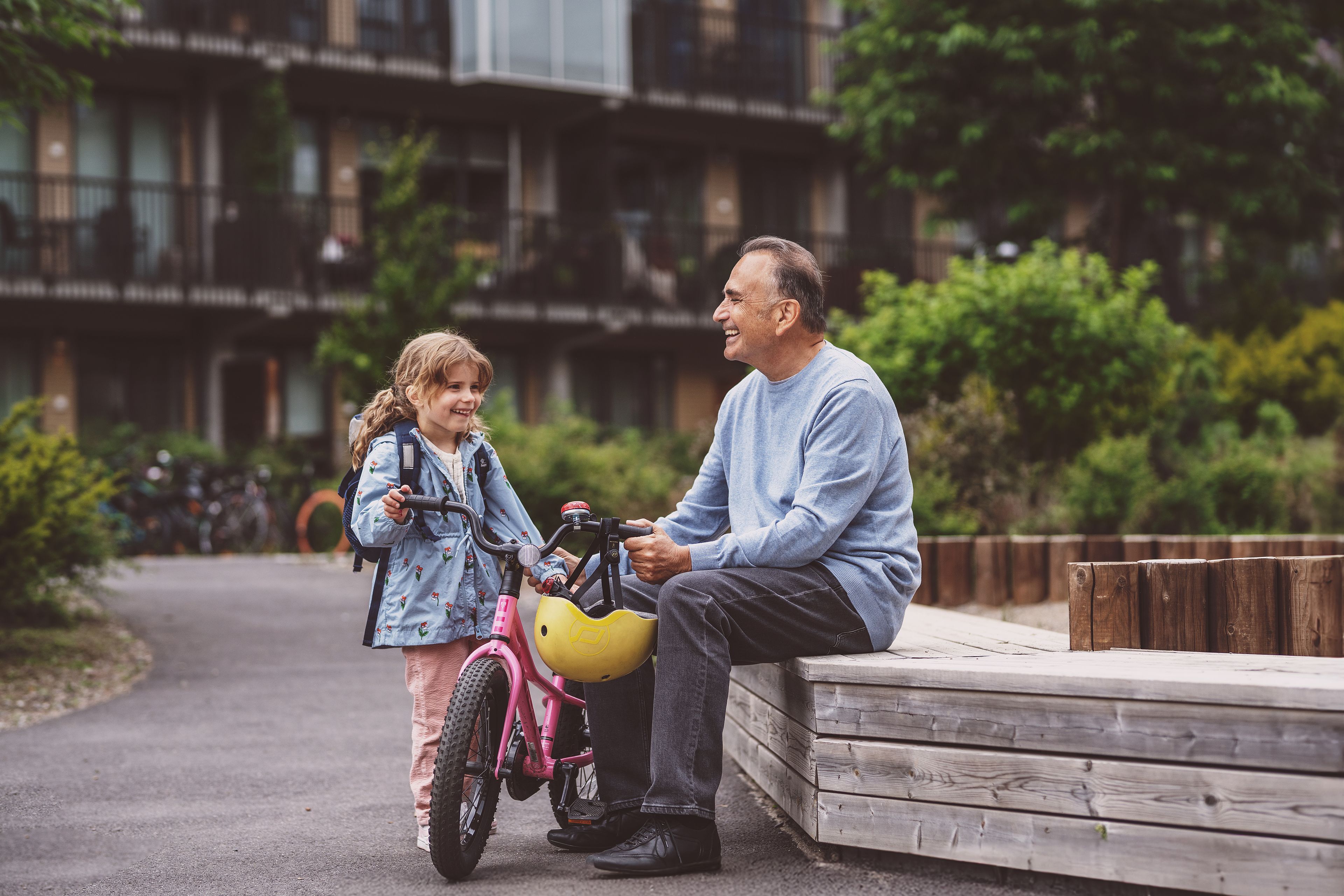 En mann sitterpå en benk og holder sykkelen til et barn. Hun står selv sykkelen, og begge to smiler.