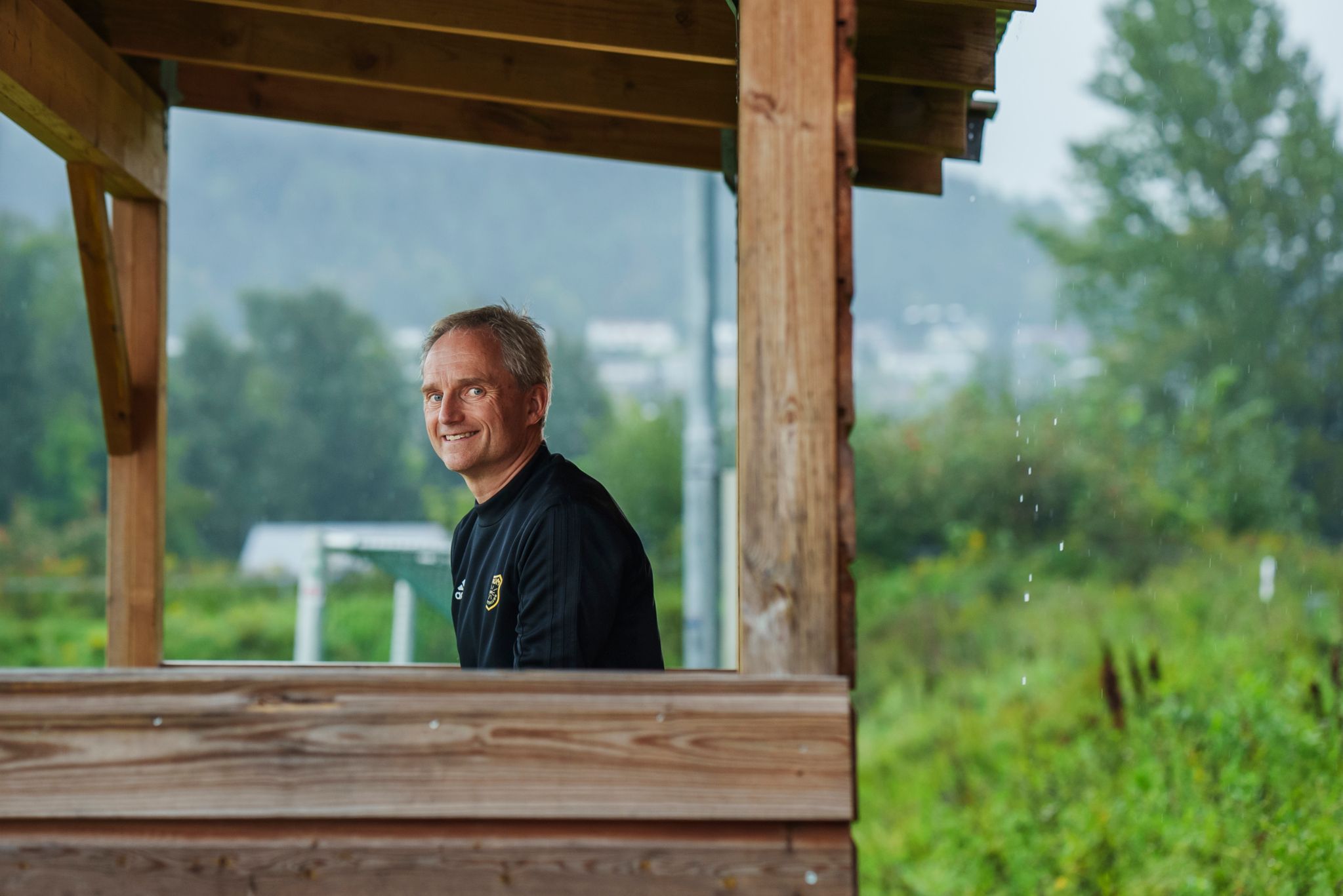 Jostein Djupvik sitter i trenerskuret og ser mot kamera, utenfor er det vått og grått.