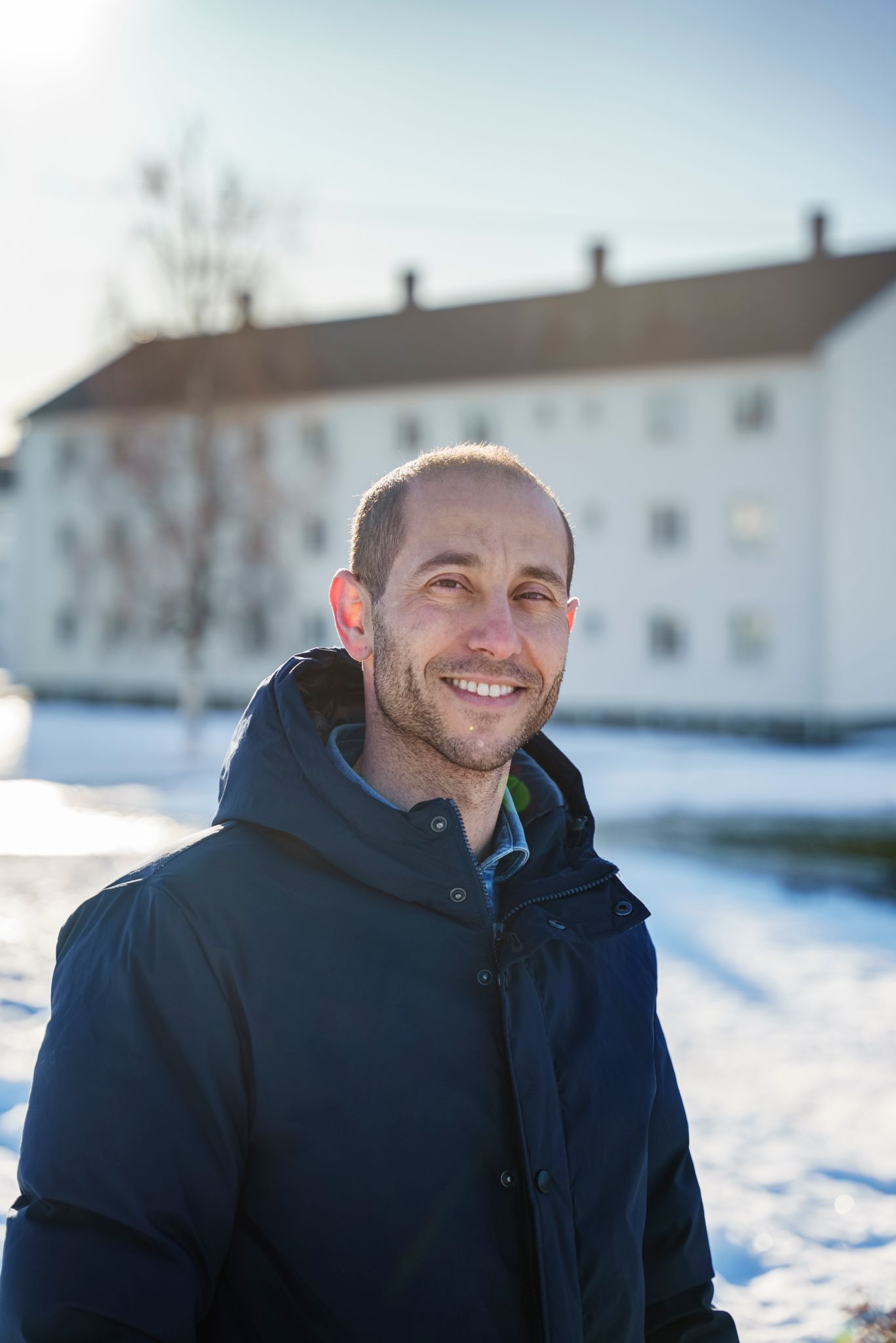 En mann står ute. Han har på seg en mørk vinterjakke. Han smiler. En blokk vises i bakgrunn.