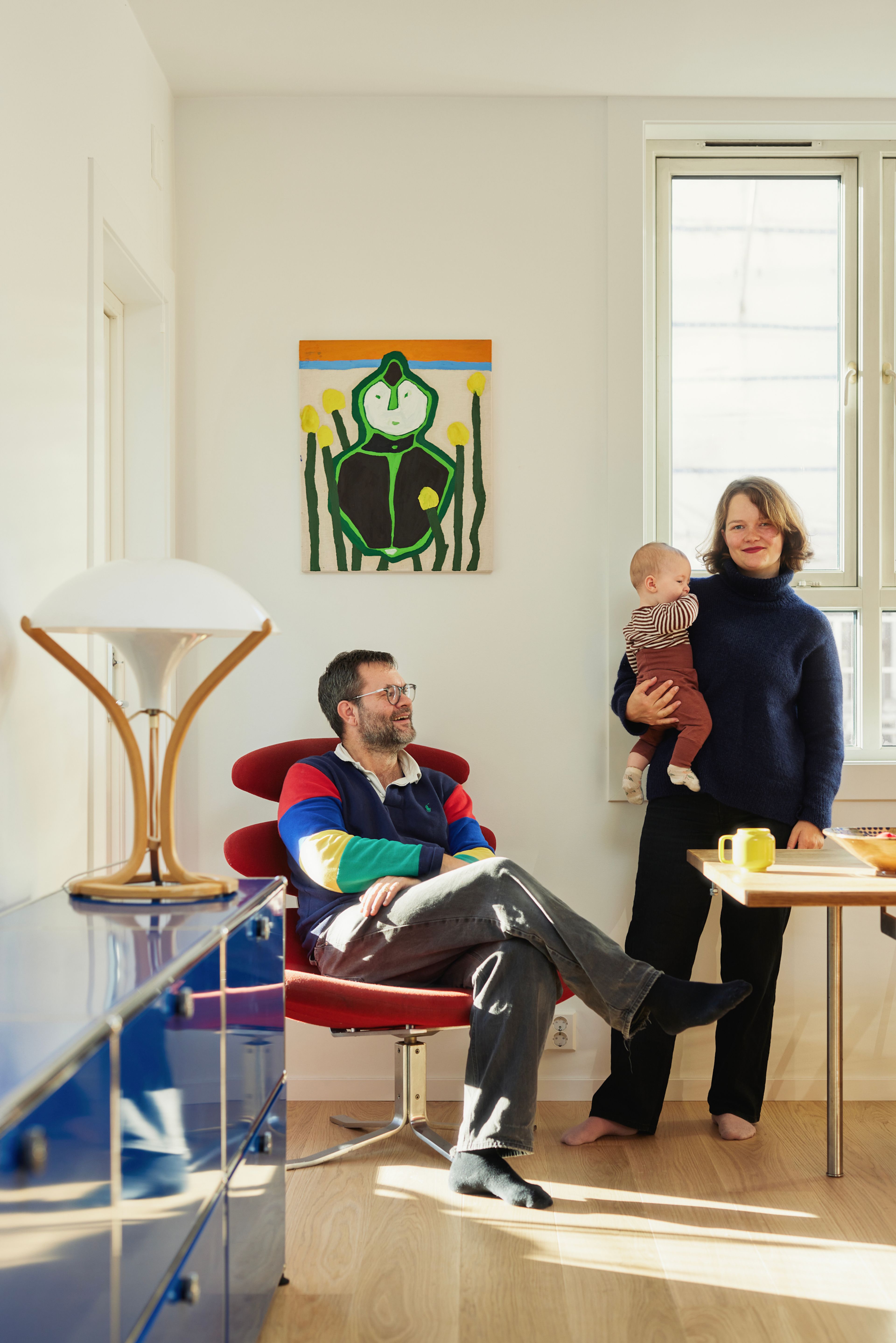 Einar og samboeren Malin er i leiligheten sin på Sagene. Einar sitter i en rød corona-stol og Malin står ved siden av med babyen på armen.