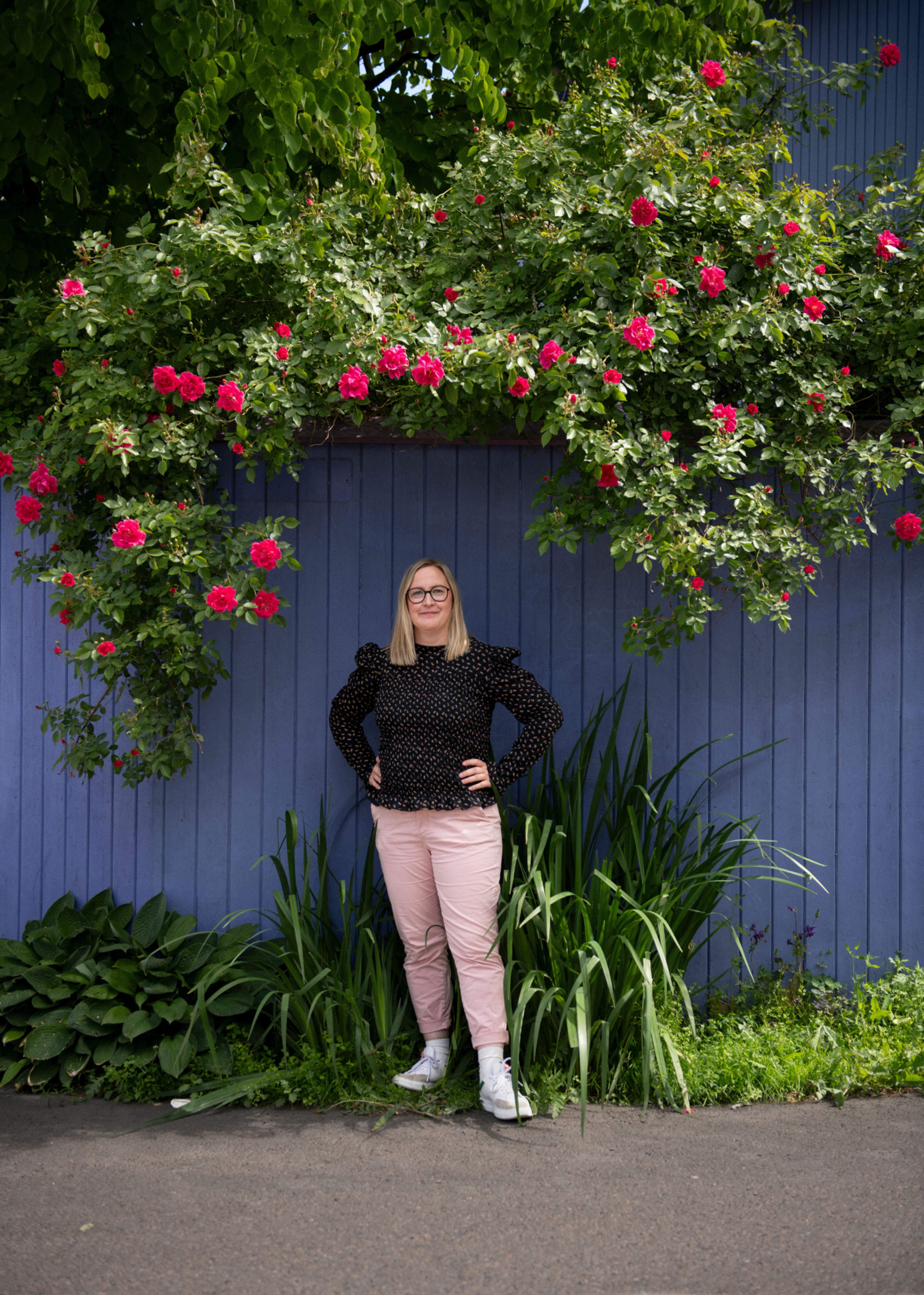 Thea Katharina Rynning står foran en blåmalt vegg og en rosebusk. Hun har på seg rosa bukse og svart topp.