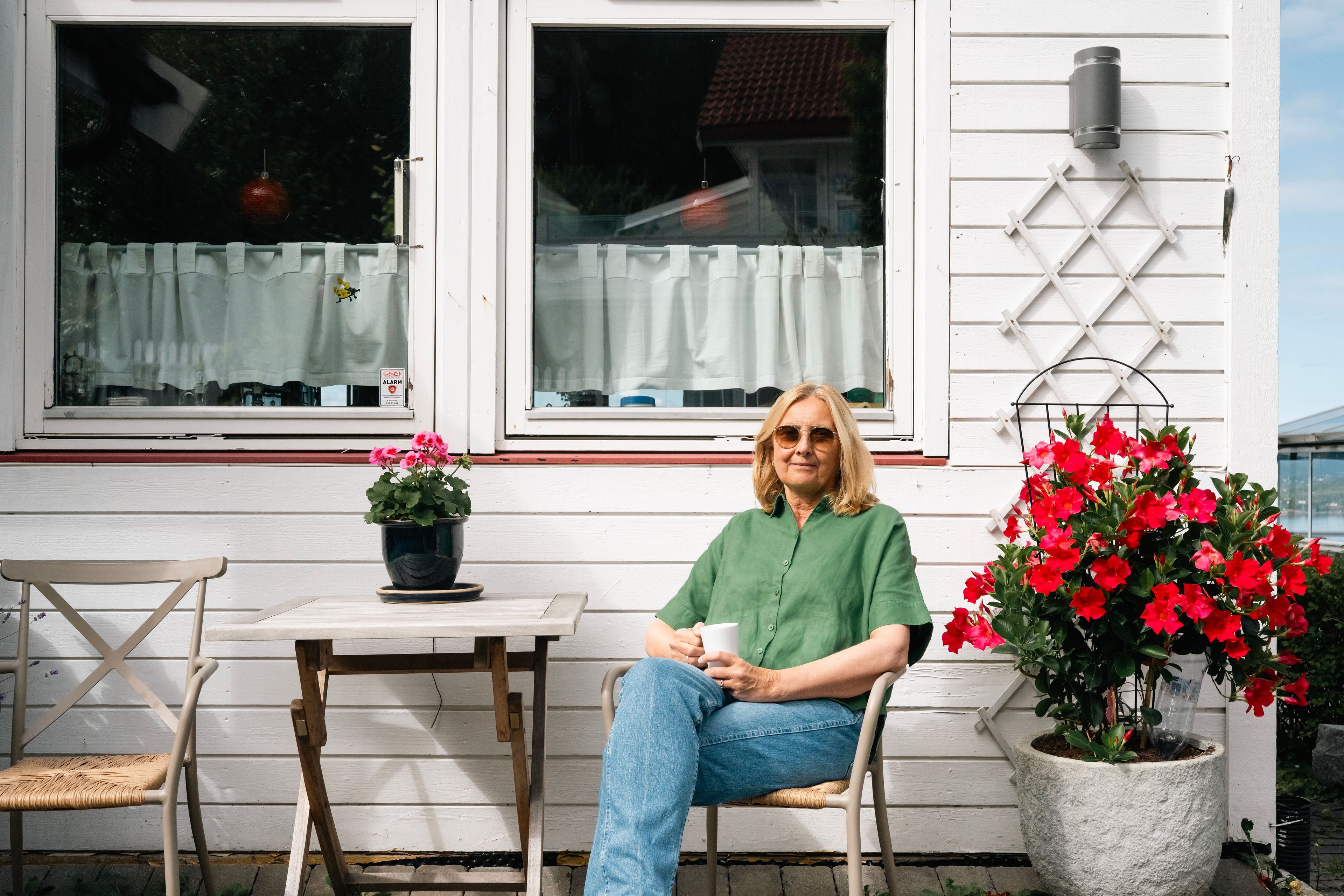 En dame sitter ved et cafebord på framsiden av et hvitt hus. Hun har på seg en grønn trøye, solbriller og drikker av en kopp.