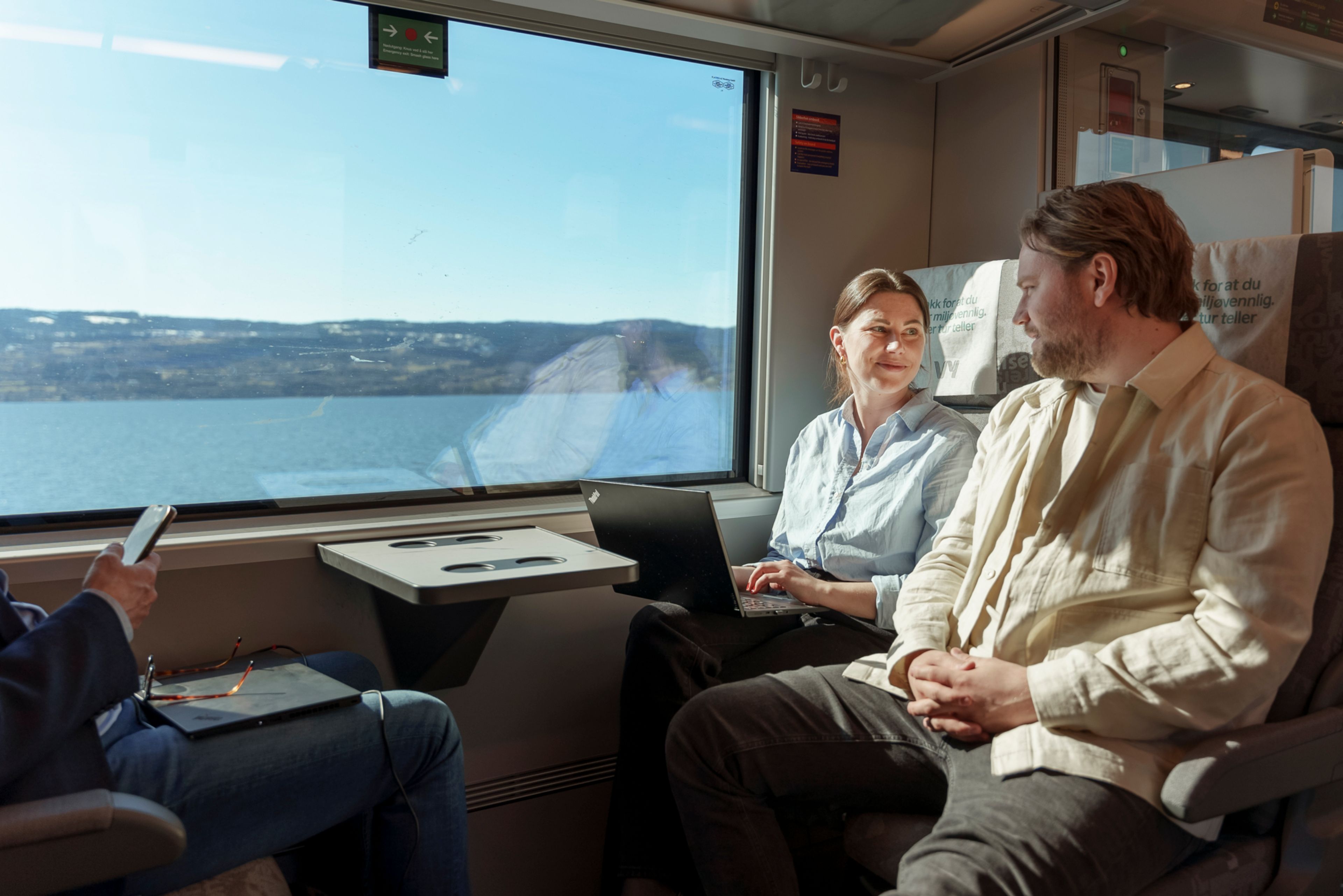 Stine og Martin sitter på toget og ser på hverandre, med utsikt over Mjøsa i bakgrunnen mot en solfylt himmel.