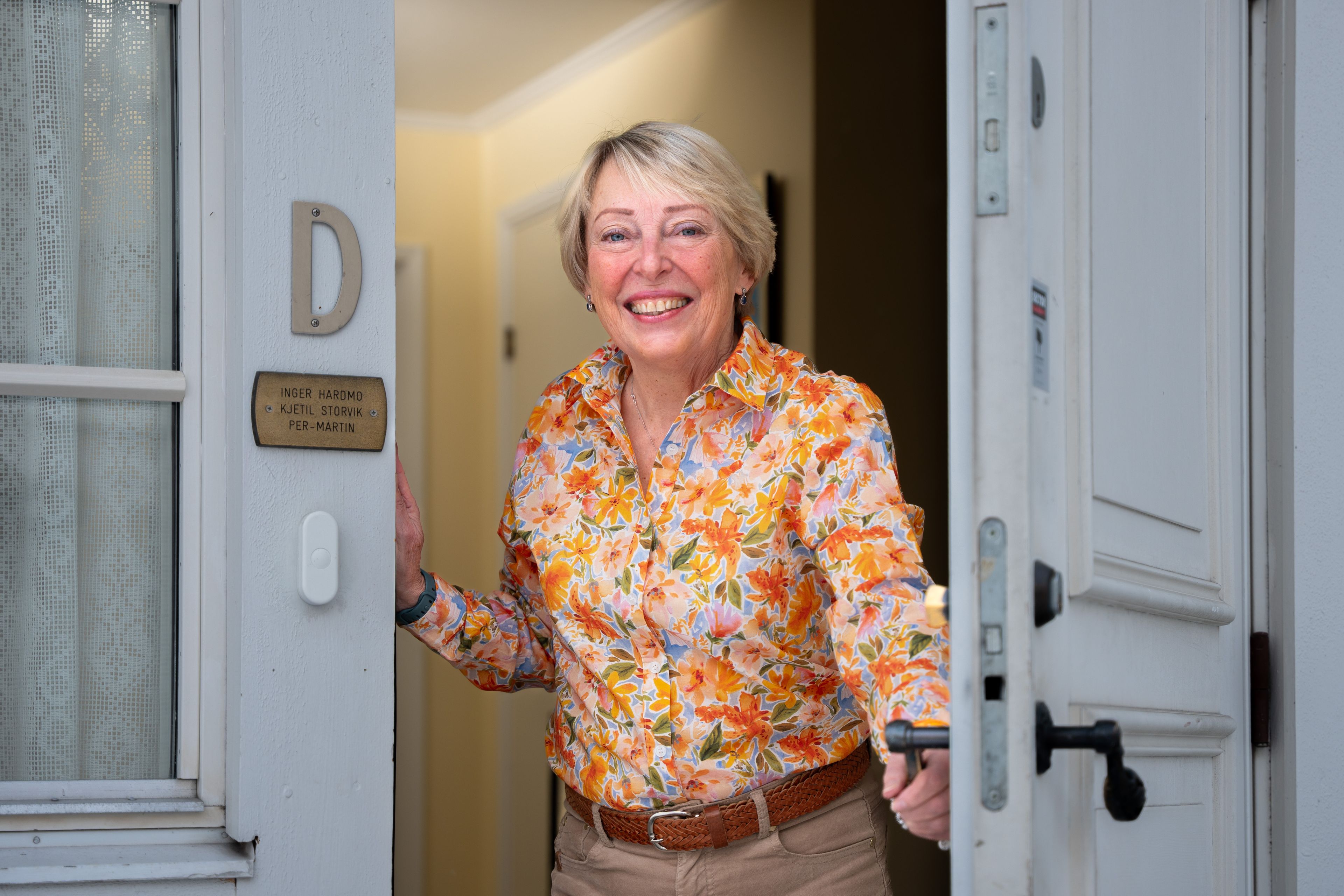 Smilende kvinne i blomstrete skjorte står i åpen inngangsdør og ønsker velkommen.