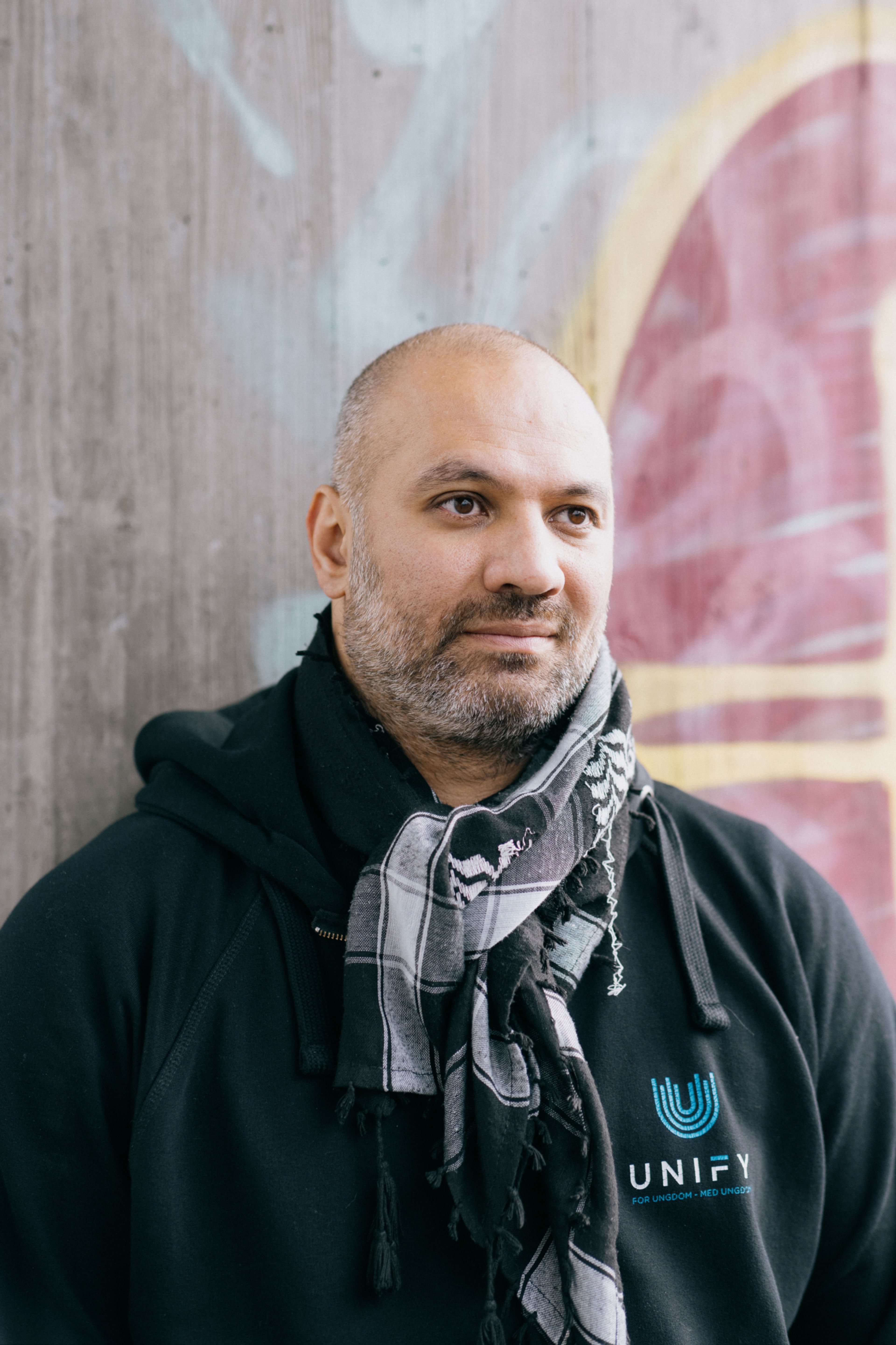 Foto av Marco Elsafadi som står foran en murvegg. Han har på seg hettegenser og palestinaskjerf.