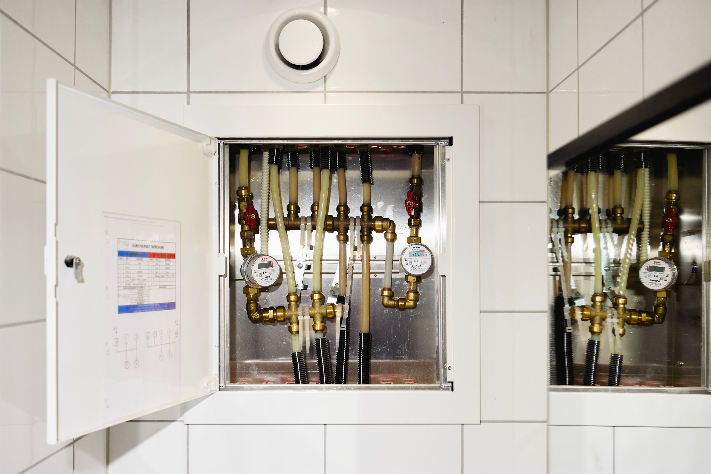 Åpent skap med rør og individuelle vannmålere på vegg med hvite fliser