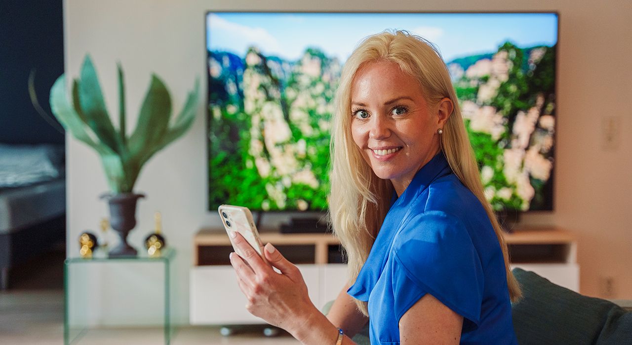 Ei dame ser mot kamera og smiler. I bakgrunnen står TVen på. Foto.
