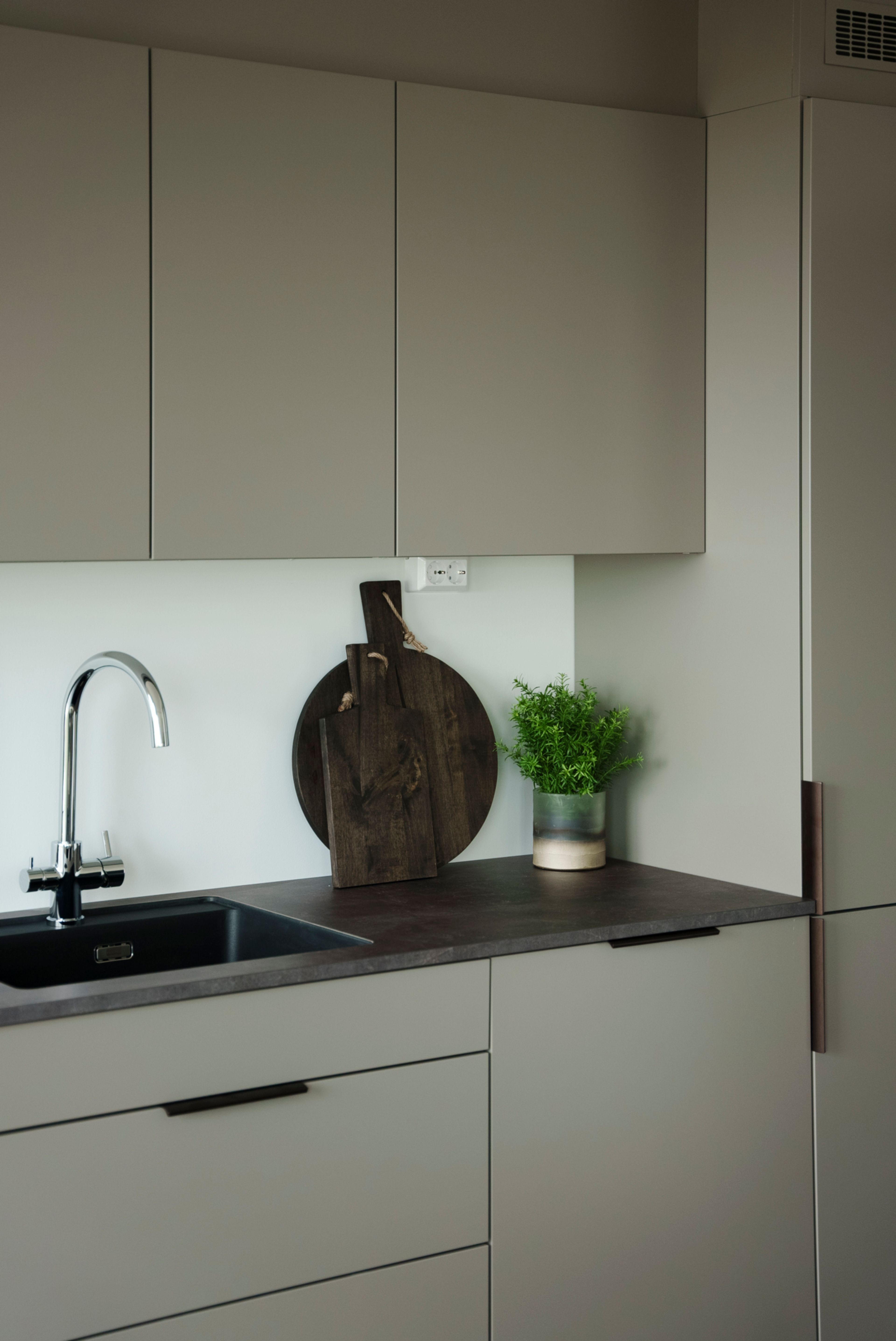 Et kjøkken med grågrønn farge på fronter, sort benkeplate og grønne planter.