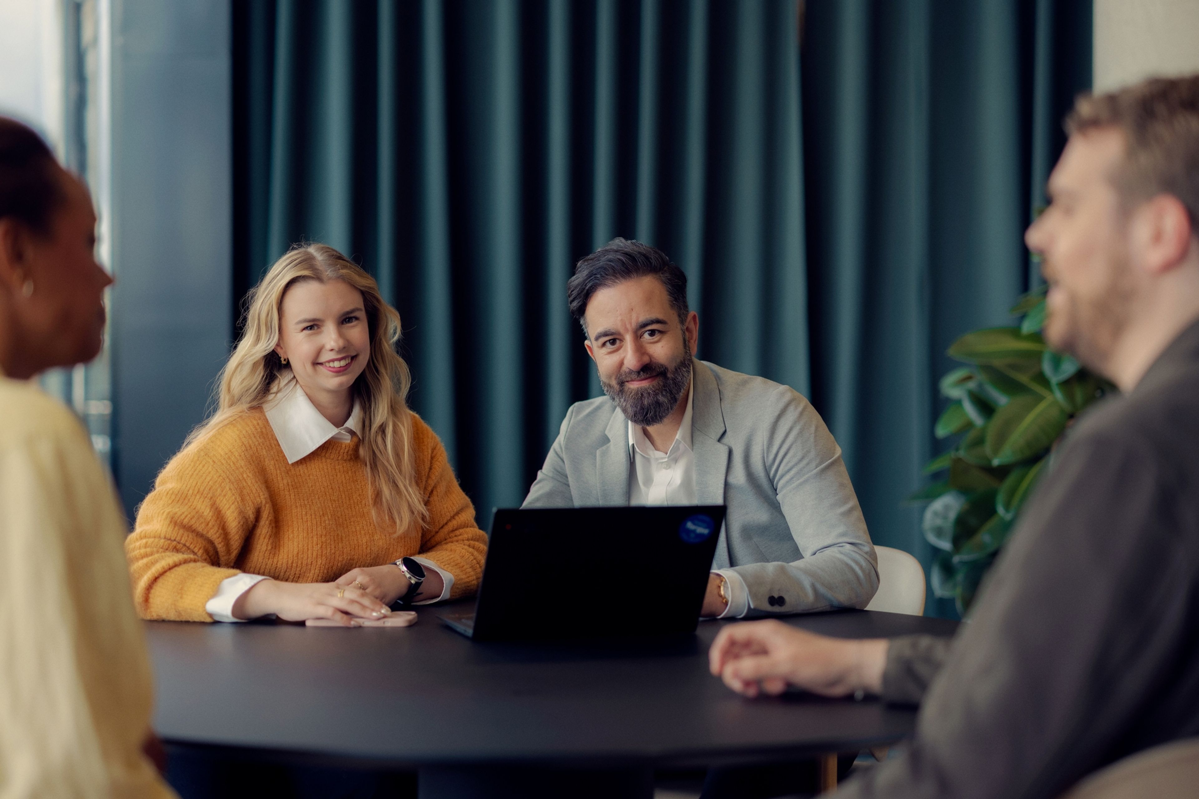 Fire personer sitter rundt et møtebord og har et møte. To av dem ser mot kamera, og en bærbar PC står på bordet mellom dem.