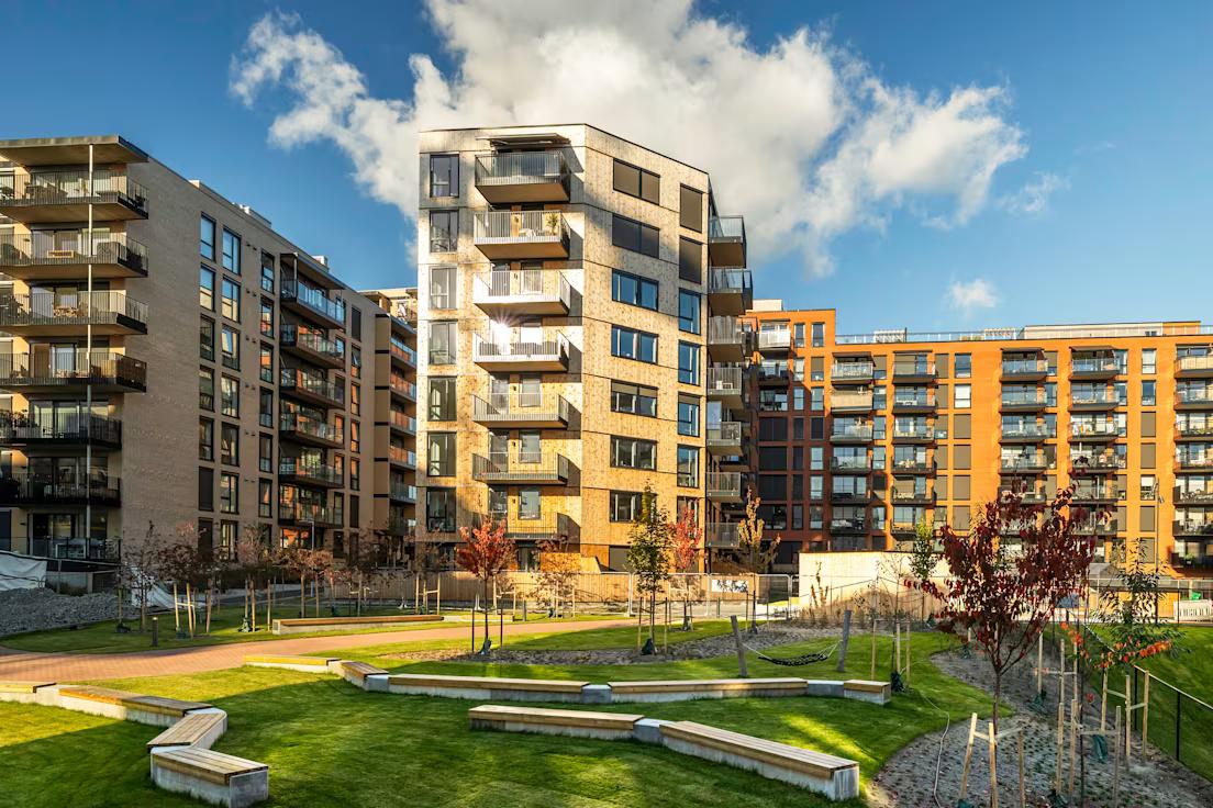 Moderne leilighetsbygg under en blå himmel med skyer. Sollys reflekteres i vinduer. En gårdsplass med grønt gress, stier og unge trær foran.
