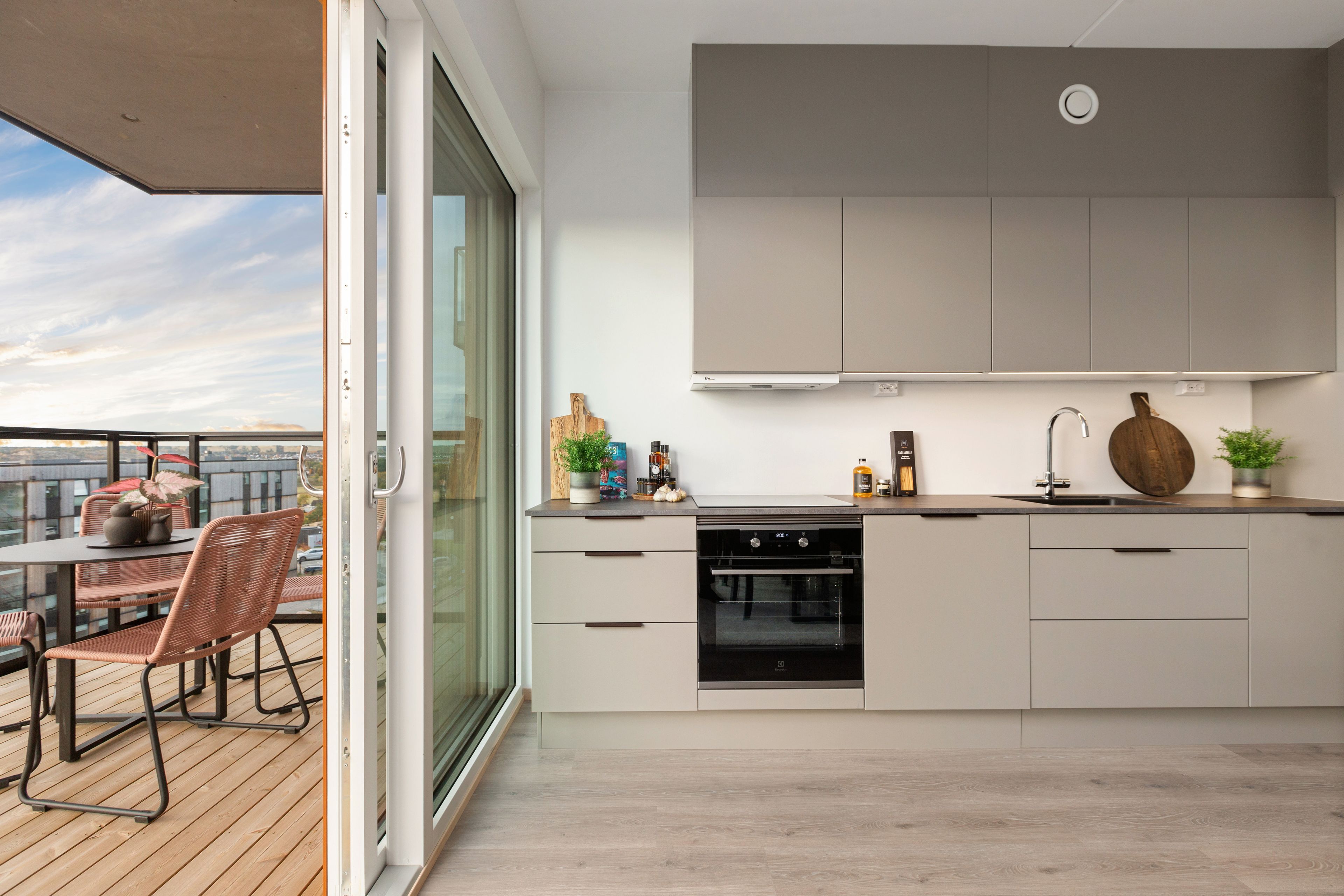 Kjøkken i en leilighet i Furukrona, med kjøkkeninnredning med beige fronter og utgang til balkong gjennom skyvedører i glass. Deler av balkongen med spiseplass er med i bildet.